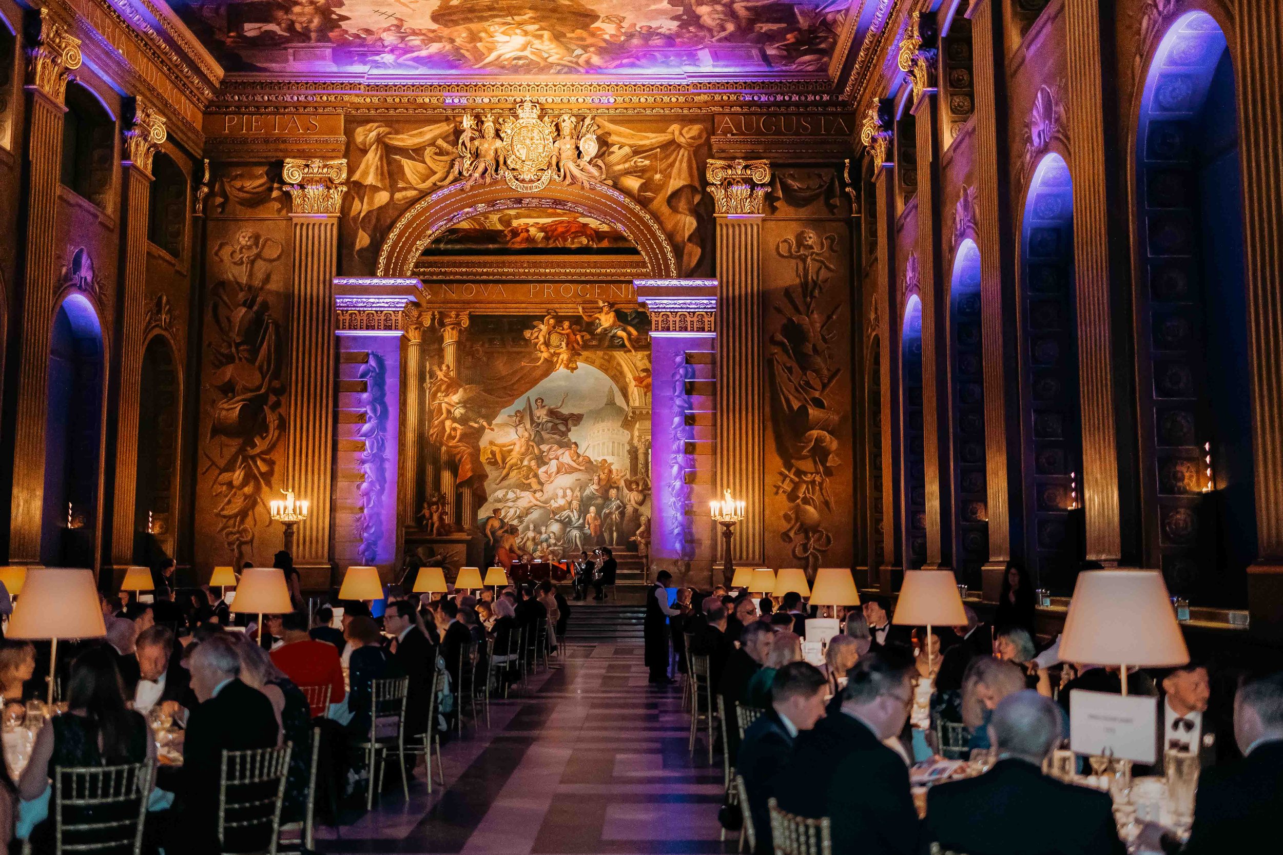 Elegant banquet hall decorated with grand paintings, ornate columns, and warm lighting. Many people in formal attire are seated at dining tables with lamps, and there is a stage with musicians arranged for a performance.