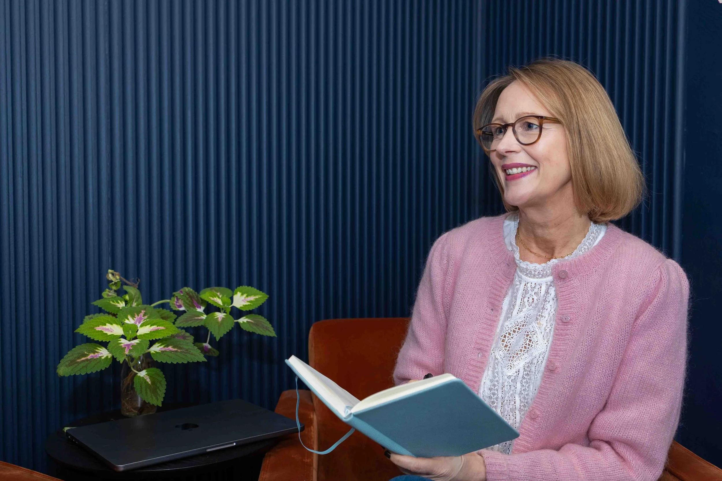 A woman with glasses and short red hair, wearing a pink sweater, sitting and smiling while holding an open notebook.