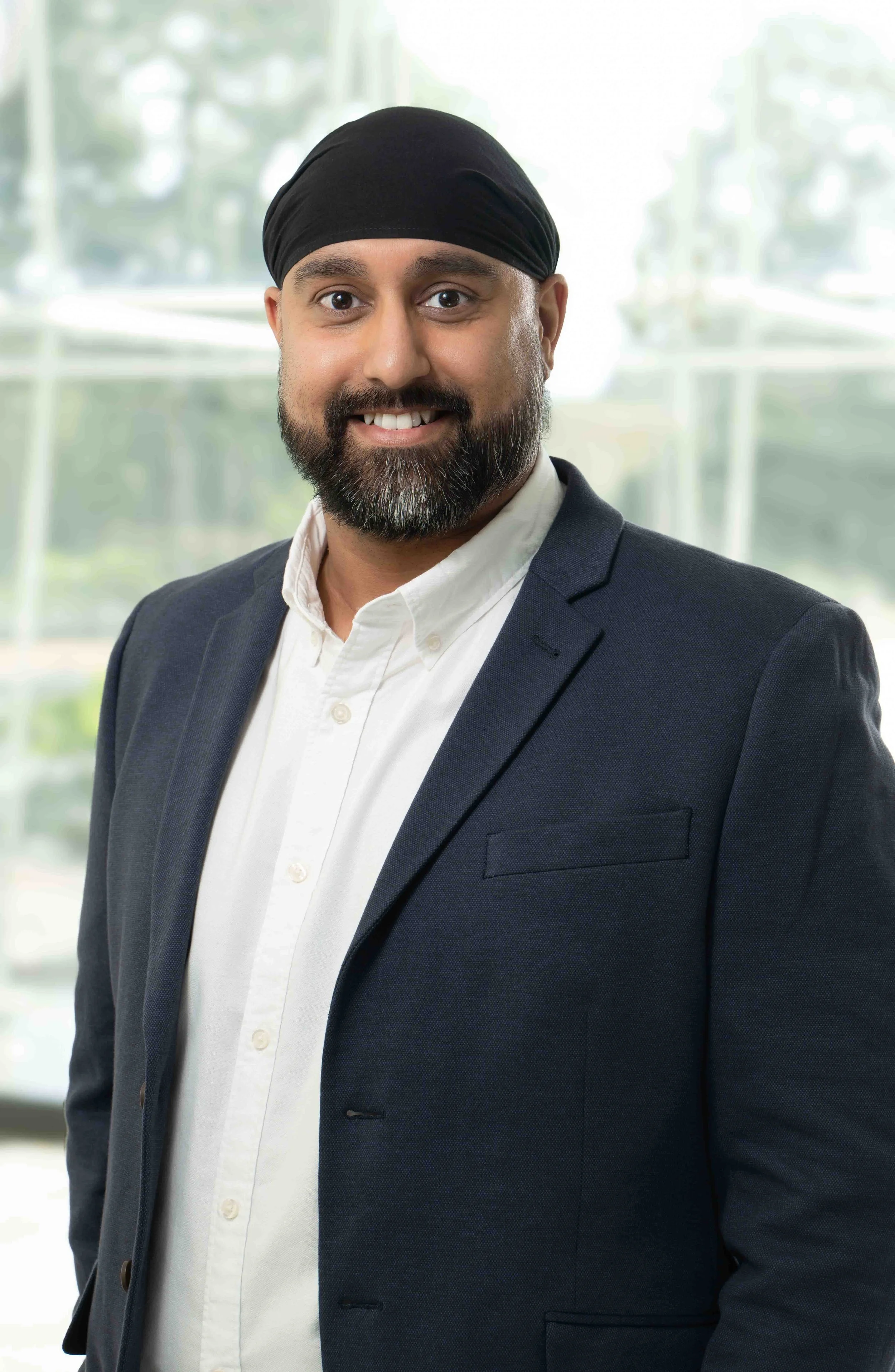 Professional man with a beard and turban smiling, wearing a navy blazer and white shirt, standing in front of a bright window.