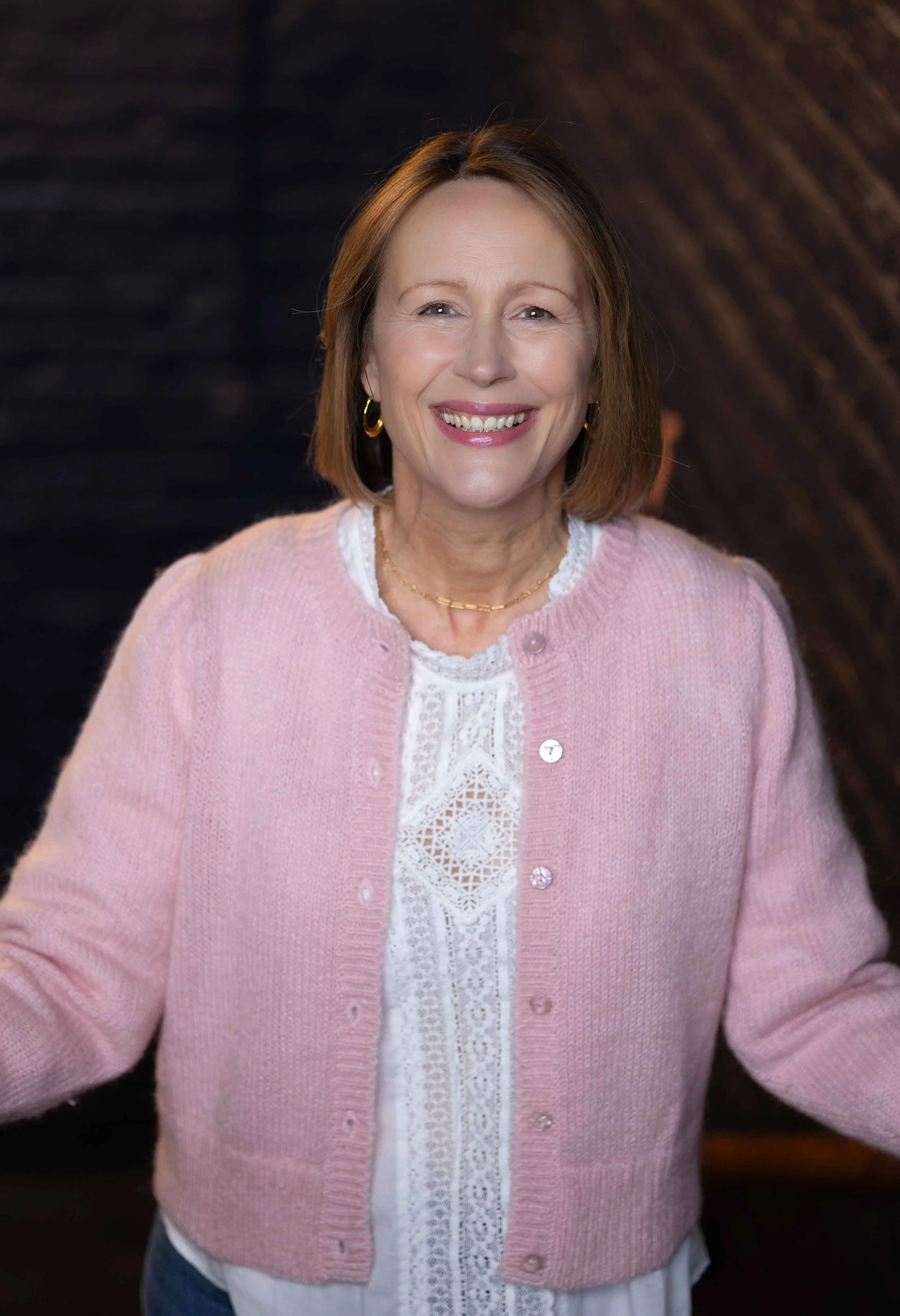 A smiling woman with shoulder-length brown hair, wearing a pink cardigan over a white lace blouse, gold hoop earrings, and a gold necklace, standing in front of a dark textured background.