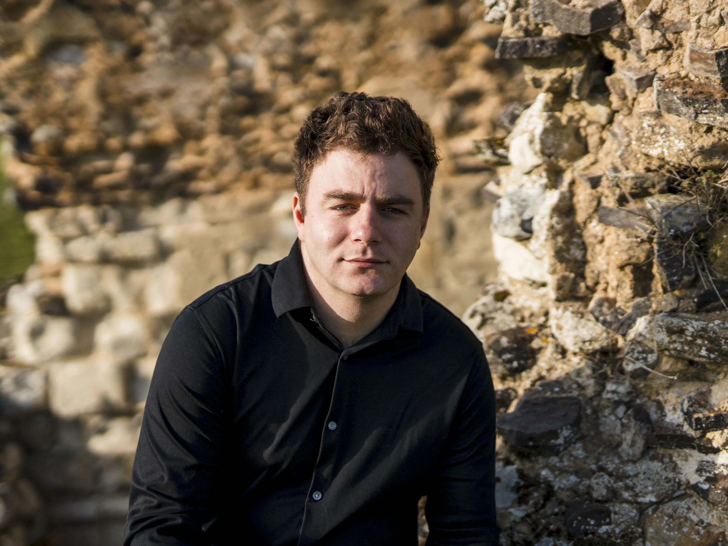 A young man with dark curly hair and fair skin, wearing a black shirt, standing outdoors against a rugged stone wall during daytime.