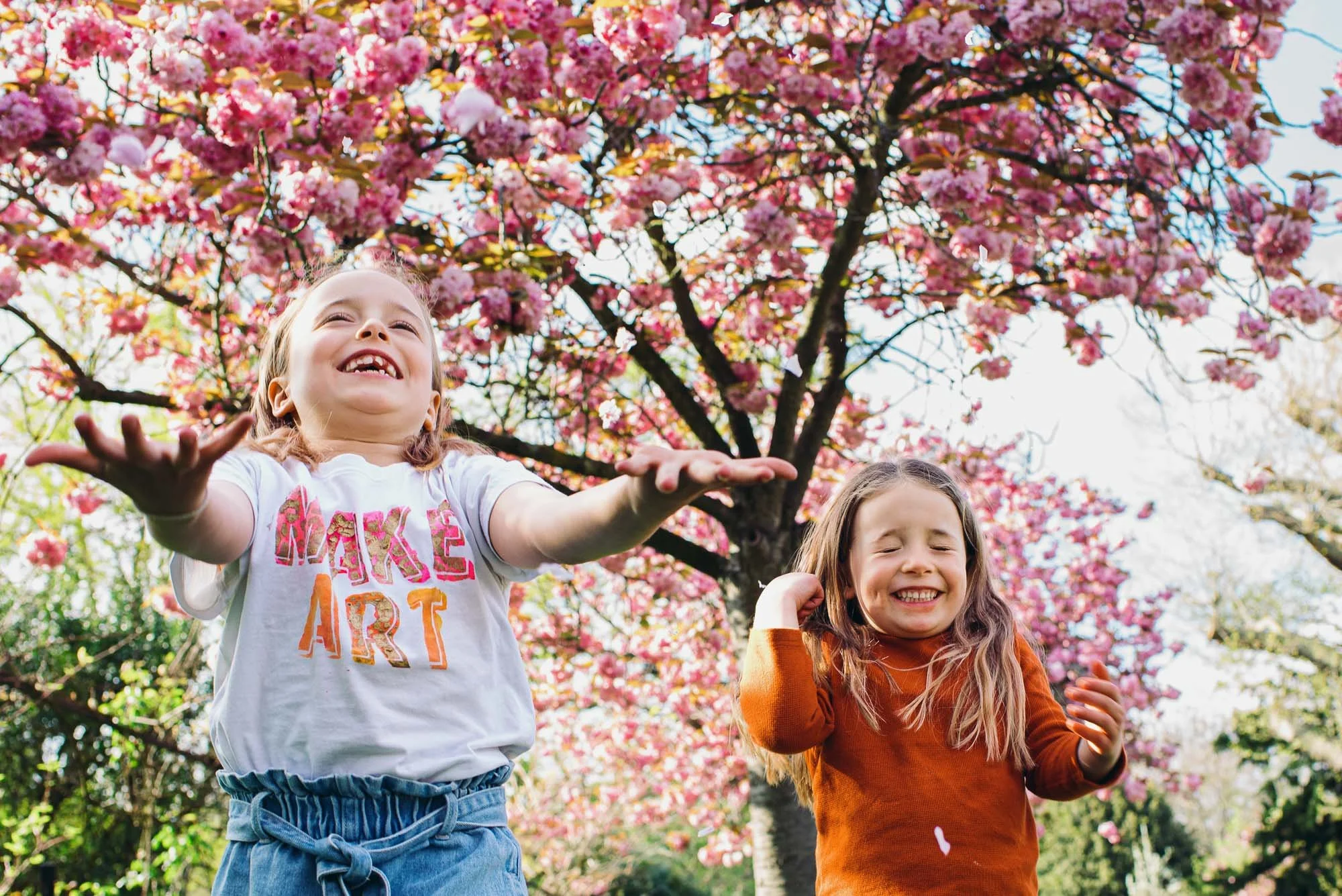 Candid unposed family photography in Chichester, West Sussex. Portrait, unpose,d of girls throwing blossom on Spring day in local park in West Sussex.