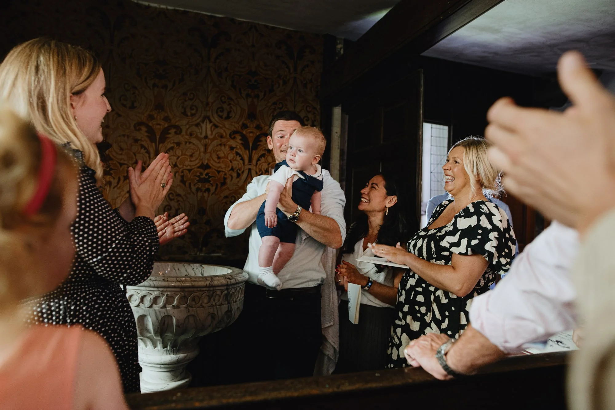 London christening photographer capturing moment after boy has been christened in a church in Kensington, London. Family clapping and laughing.