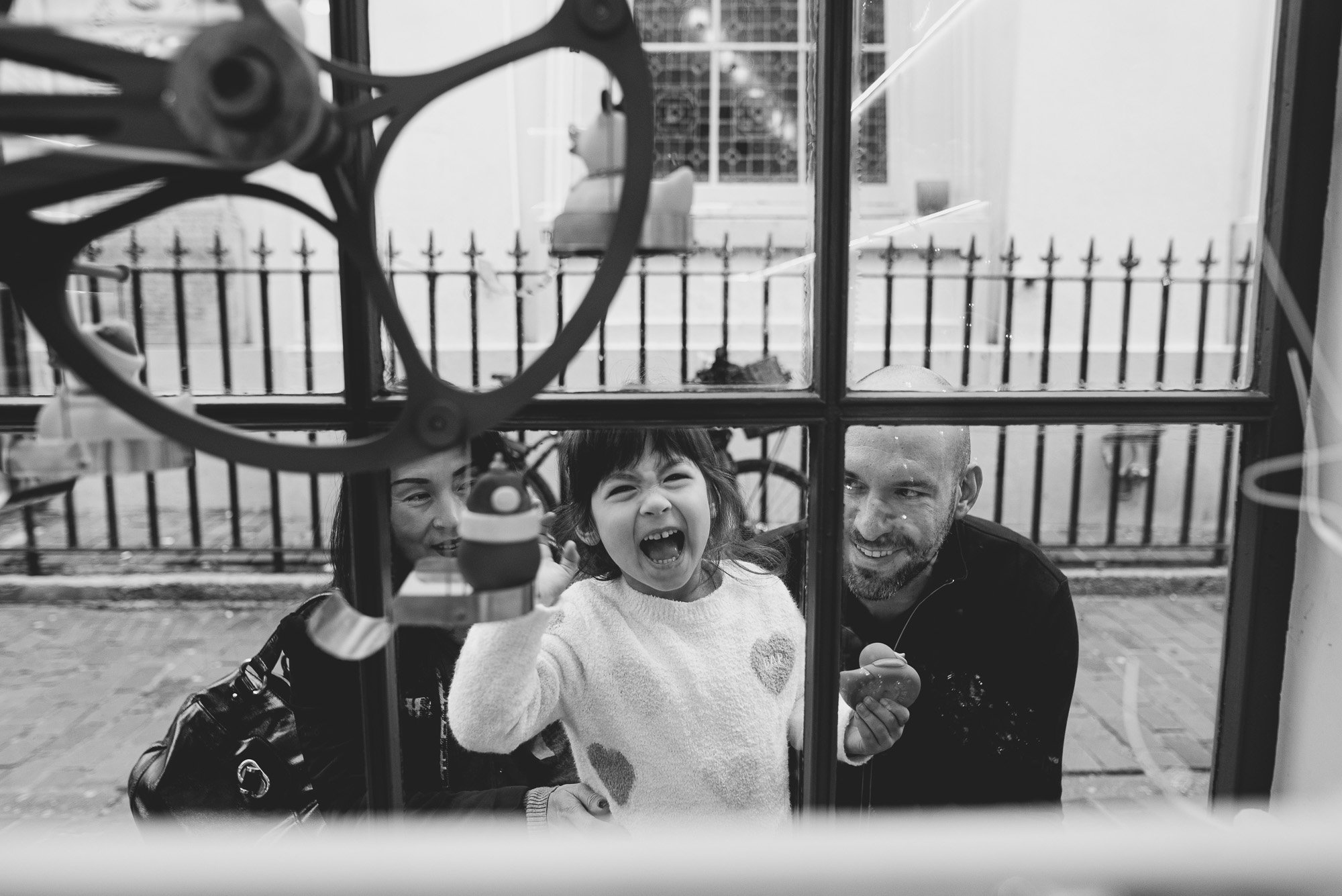 family-portrait-taken-through-shop-window-brighton-sussex-documentary-family-photos-laughing-girl-parents-sussex-family-photographer.jpg