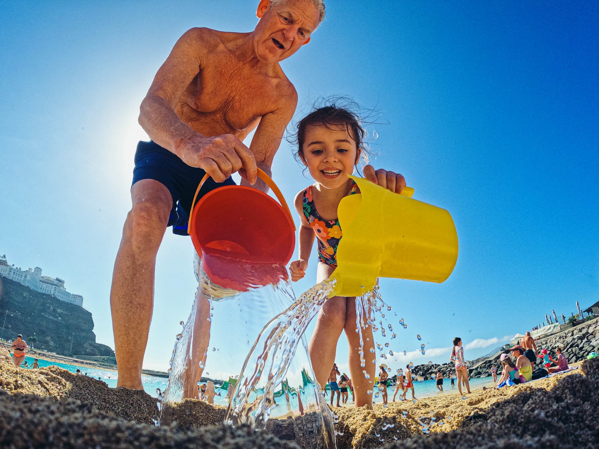girl-granddad-pouring-water-colourful-beach-buckets-summer-day-unposed-family-photography-photojournalism-documentary-approach.jpg