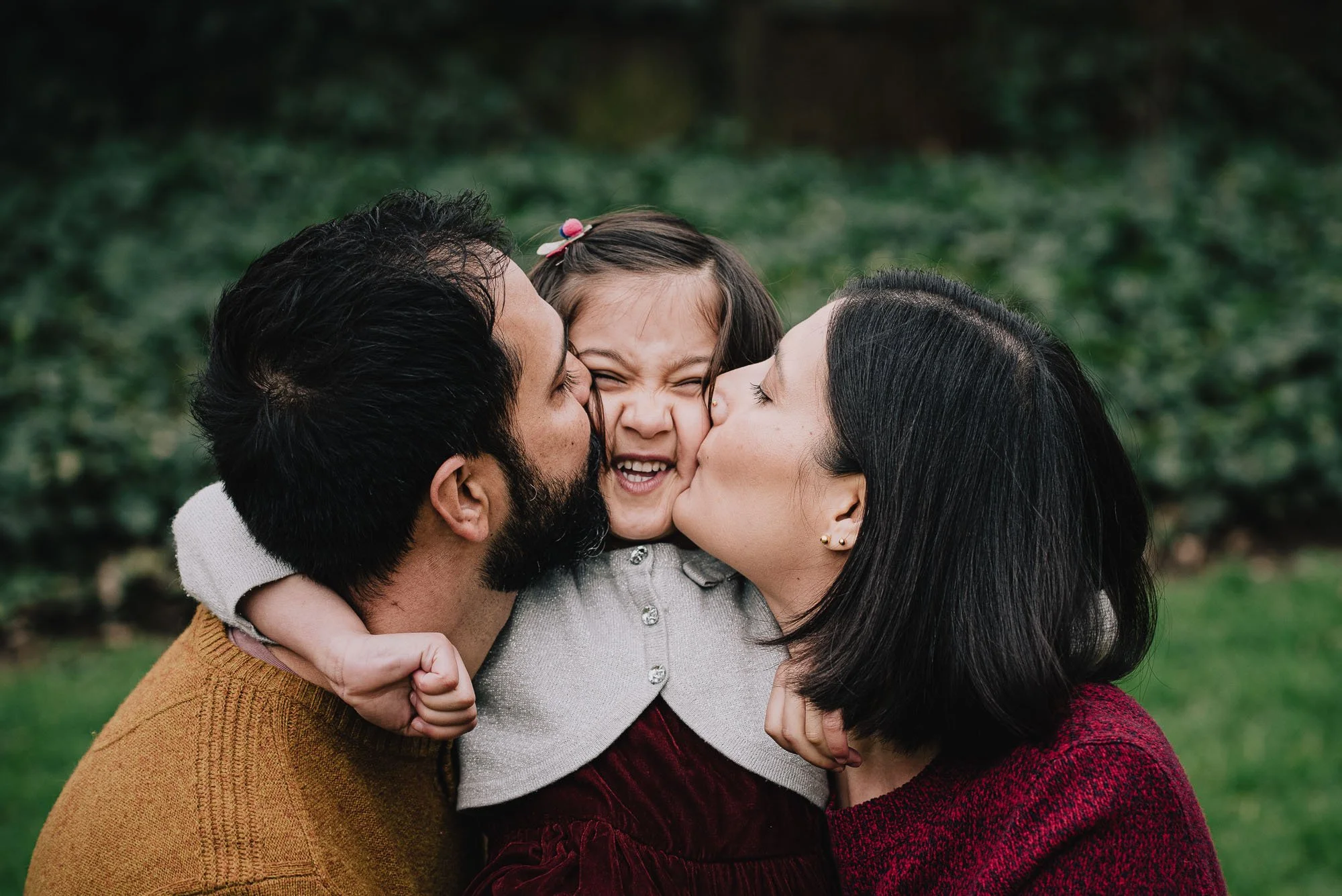 Streatham family photographer capturing daughter being kissed by both pf her parents during an outdoor London family photoshoot on Streatham Common.