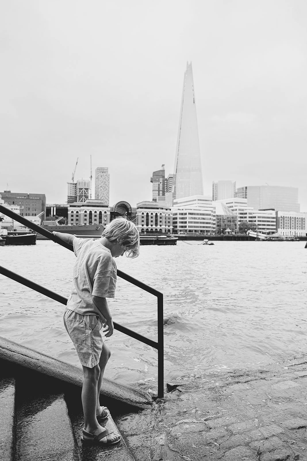 Boy on stairs near the Thames, the Shard in the background. Black and white documentary style family vacation photography in London.
