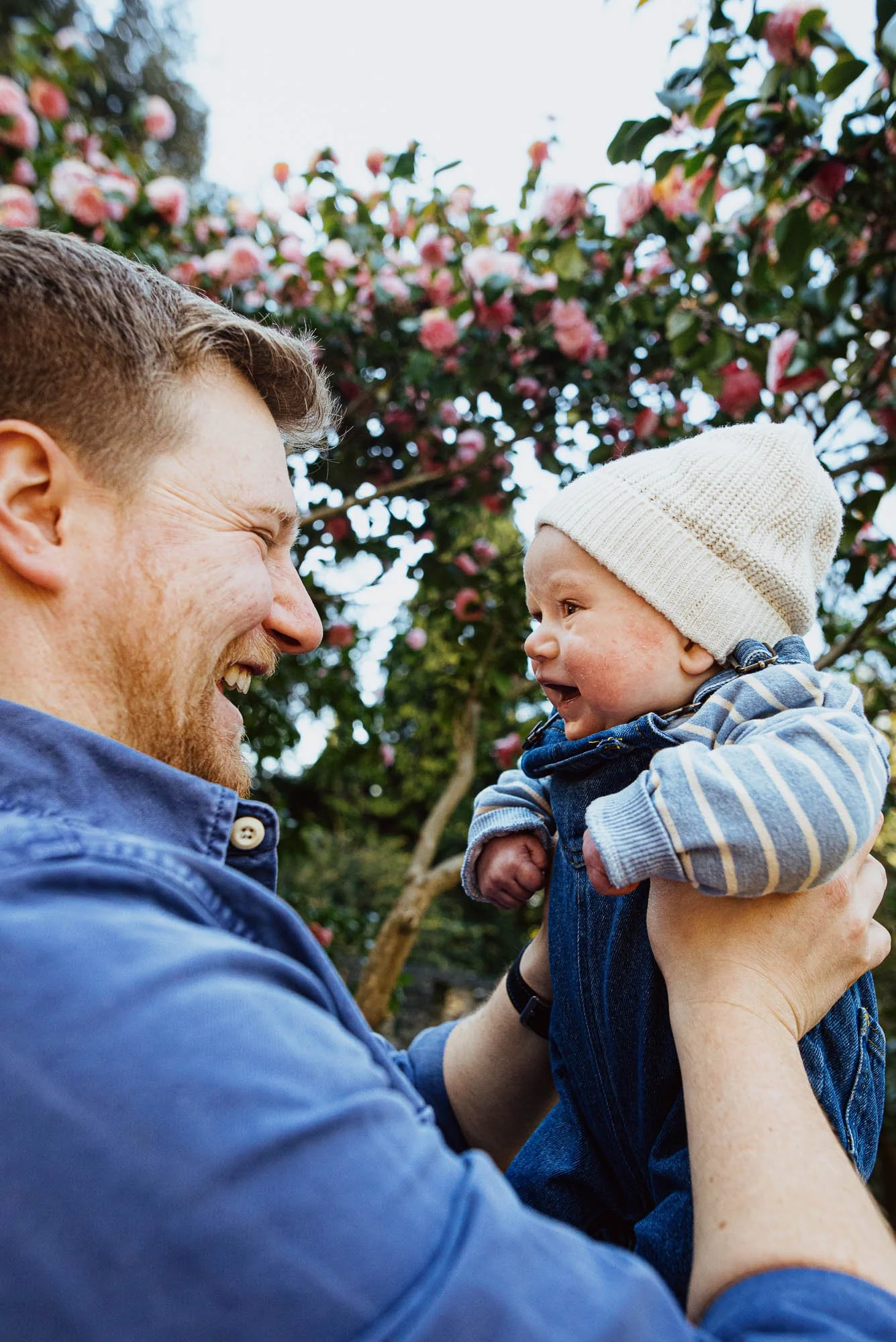 Park family photoshoot in Cannizaro Park in Wimbledon, South West London. Photo of dad and newborn baby boy, blossom in background. Unposed reportage-style family photography.