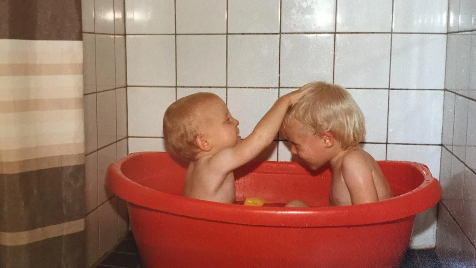 Two young children with blond hair playing together in a red plastic bathtub in a tiled bathroom. One child is touching the other's forehead while they both smile. Sussex family photography.