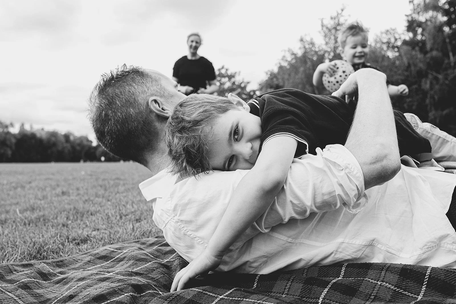Clapham family photographer pricing brochure. Dad and son hugging on Clapham Common, brother and mother in the background playing football.
