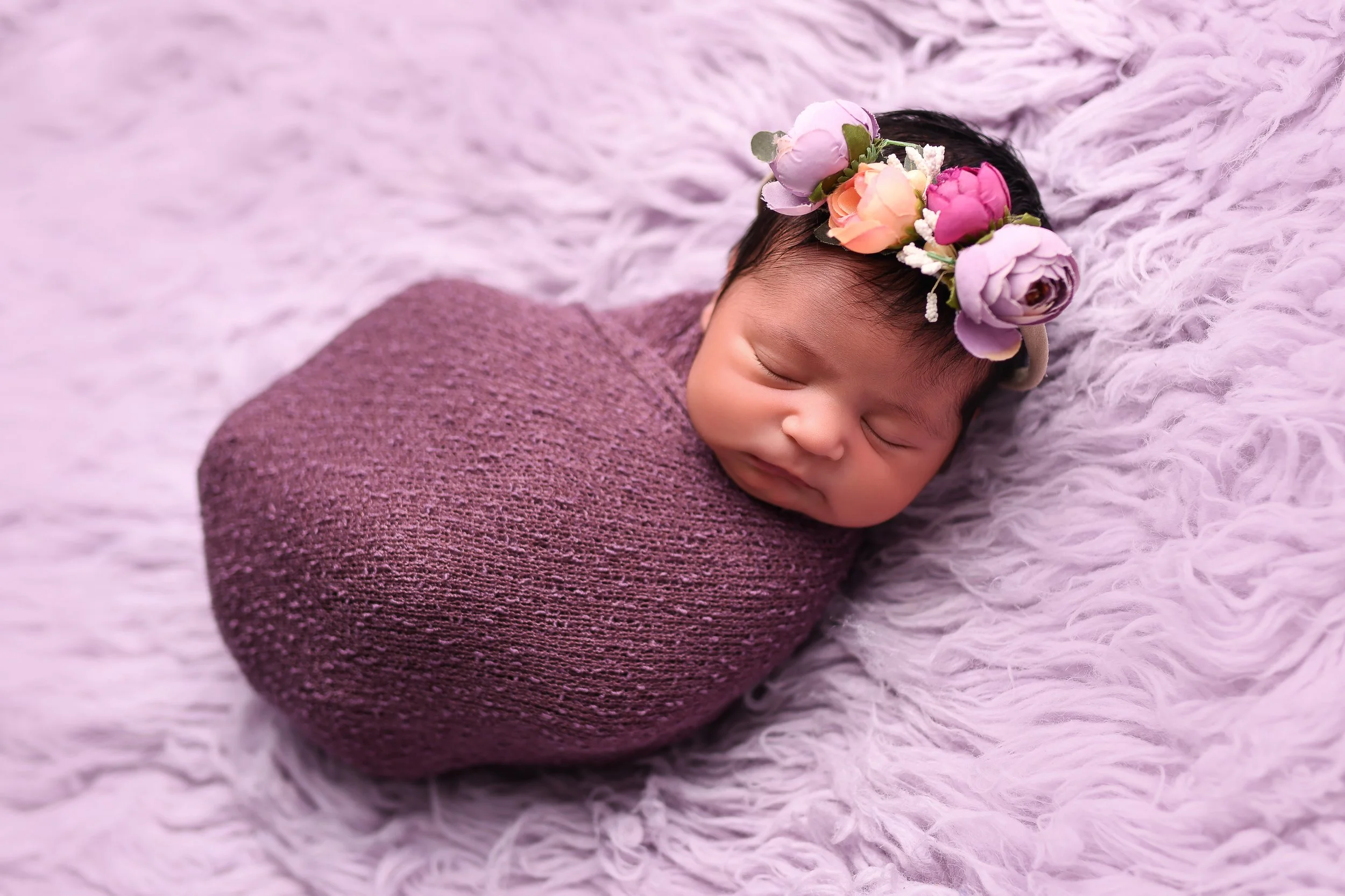 A sleeping newborn baby wrapped in a purple knitted blanket on a soft pink furry surface, wearing a floral headband with pink, purple, and peach flowers.