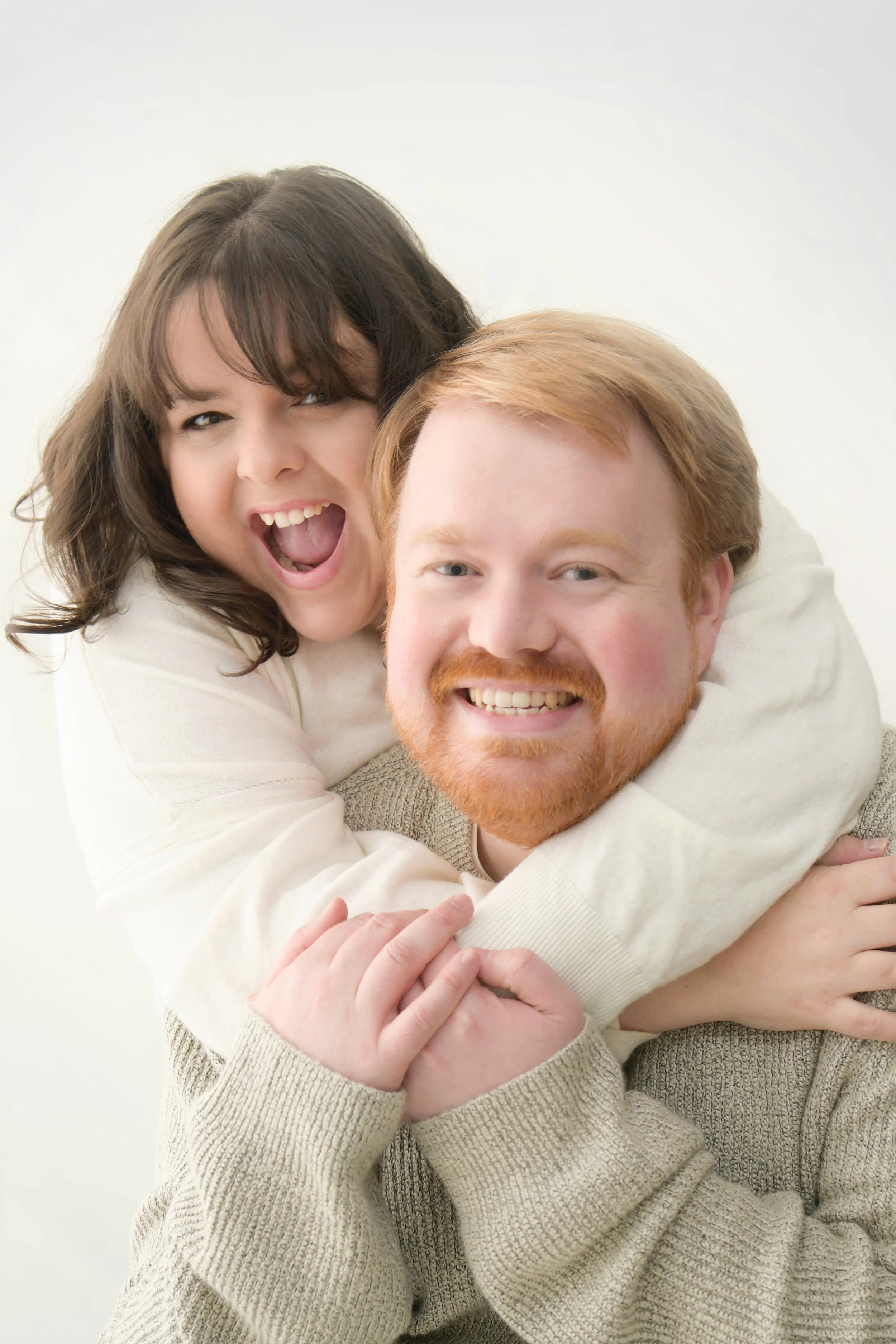 A smiling woman with brown hair and a man with red hair and beard, both embracing and looking at the camera against a plain light background.