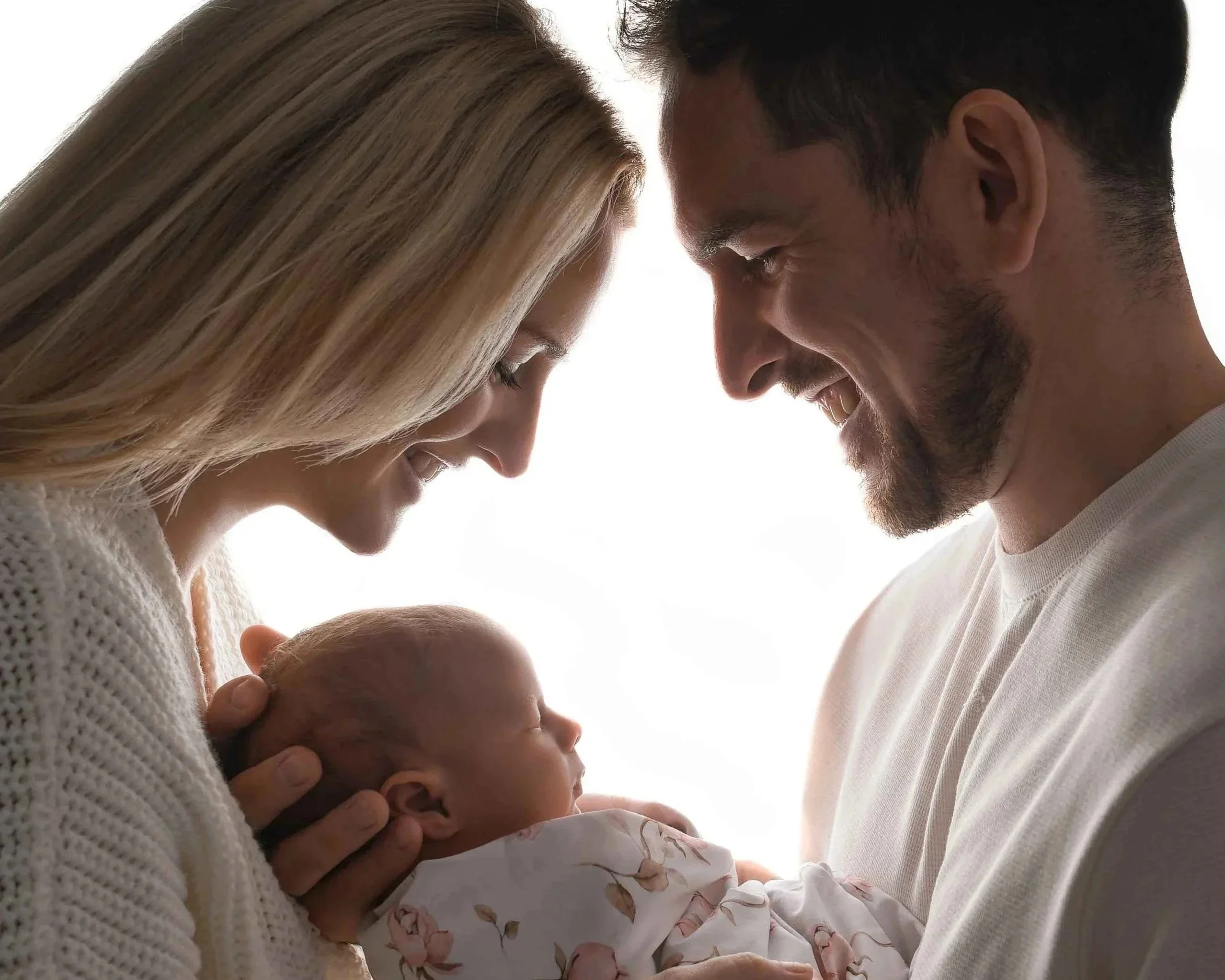 A man and a woman with light skin holding a baby close, faces touching, smiling gently, against a bright background.