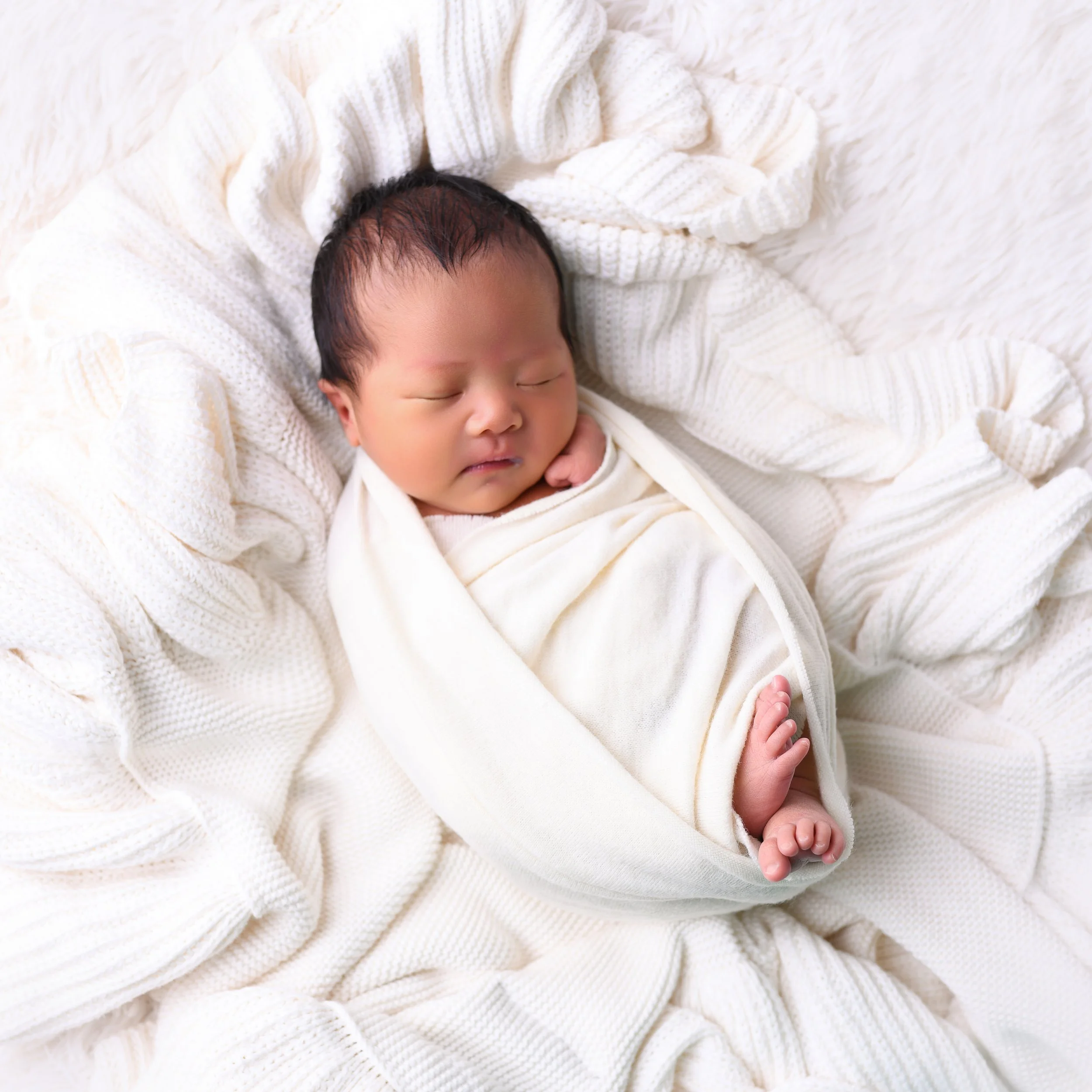 A sleeping newborn wrapped in a white blanket, lying on a soft, cream-colored knitted blanket.