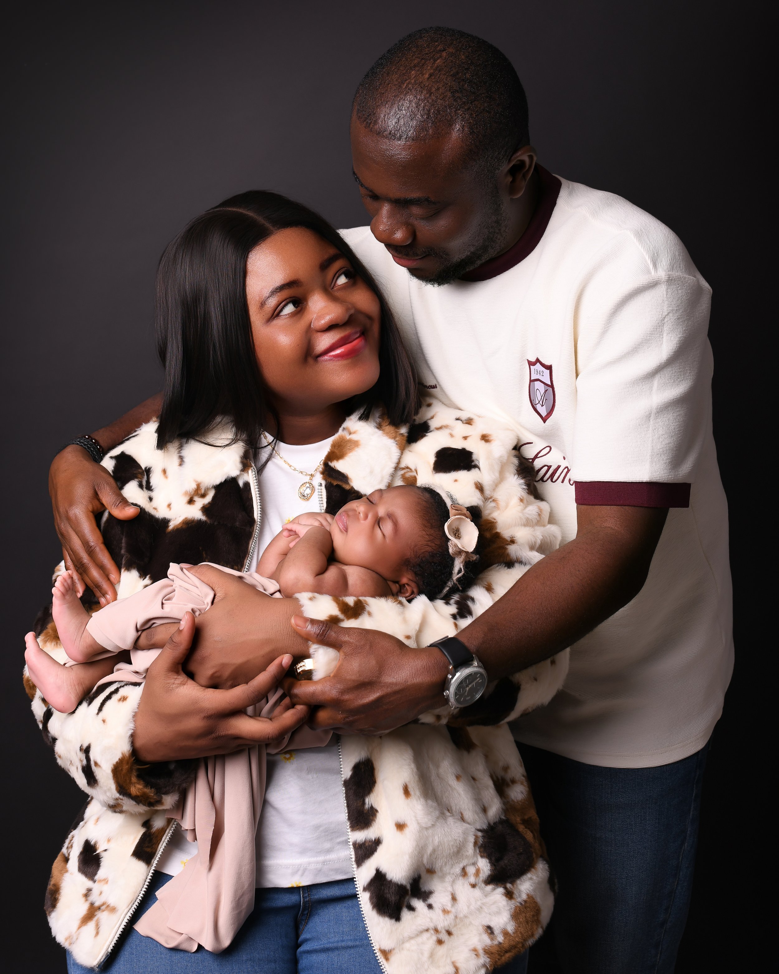A family with a woman, man, and newborn baby poses together against a dark background.