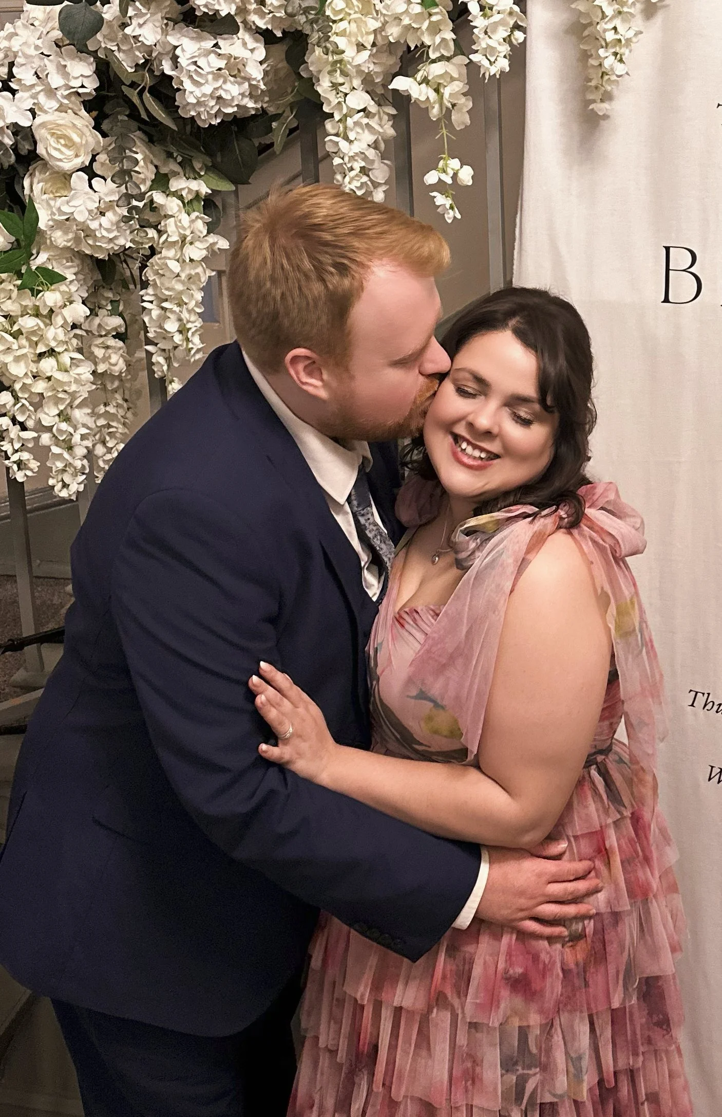 Get to know your photographer - Cara is poses with her husband. They are hugging each other, smiling, and dressed in formal attire (at a wedding). The background features white flowers and greenery, indicating a wedding or special event.