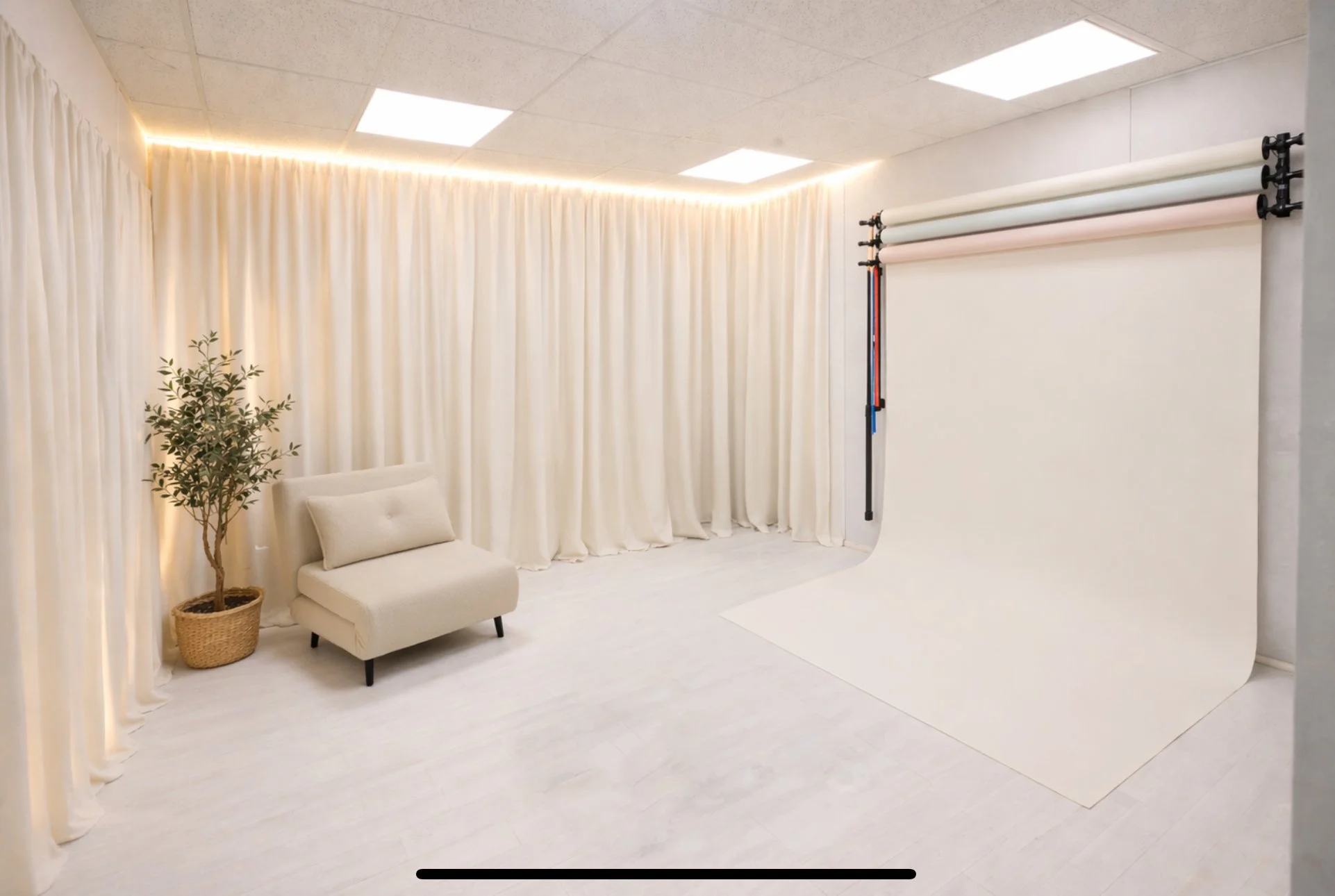 Warwickshire newborn studio plans - Photo of a cosy photography studio with white curtains, neutral seating with a pillow, a potted plant, and a photo backdrop roll on the stand.
