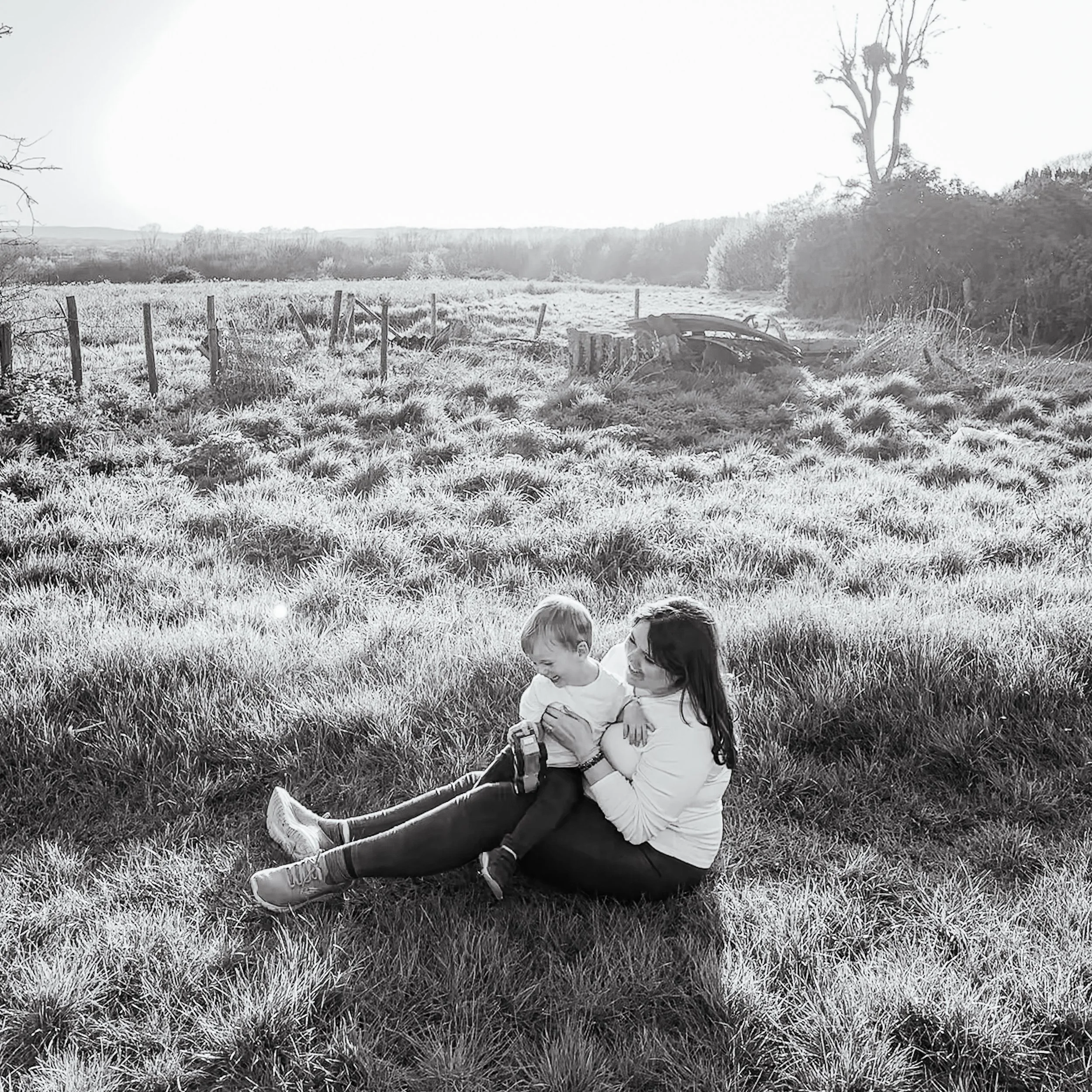 A woman and a young boy sitting together on grass in a field, smiling and enjoying each other's company, with trees and a fence in the background.