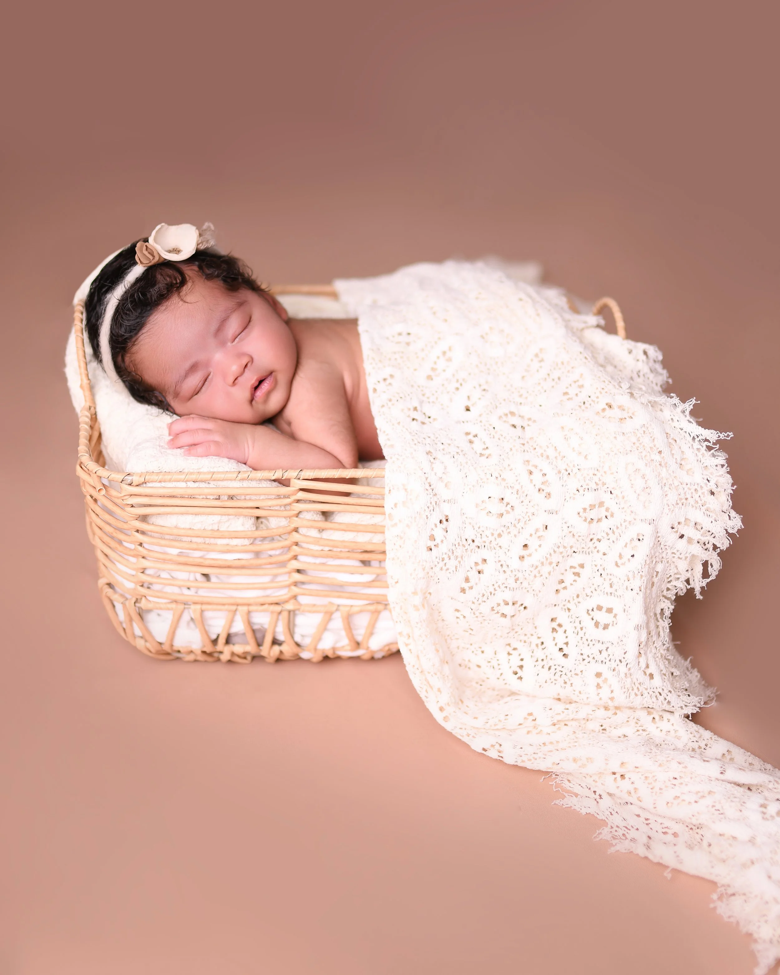 A sleeping baby lying in a wicker basket, covered with a lacy white blanket, with a headband featuring a bow on her head, on a solid brown background.