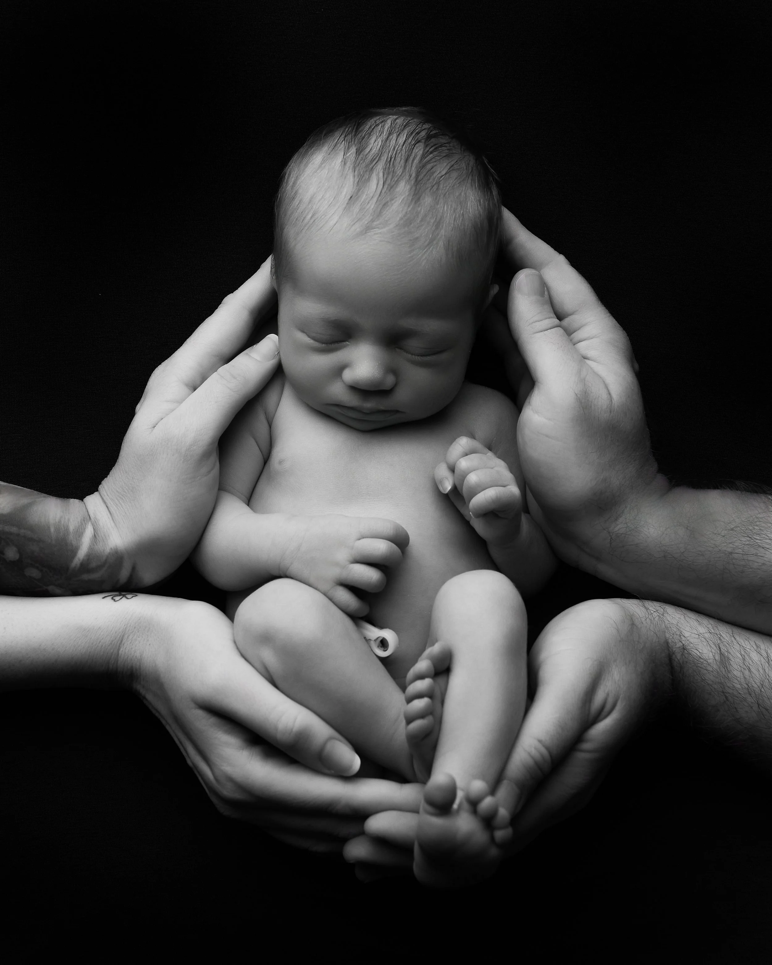 Black and white photo of a sleeping newborn baby held gently by multiple adult hands, with a dark background.