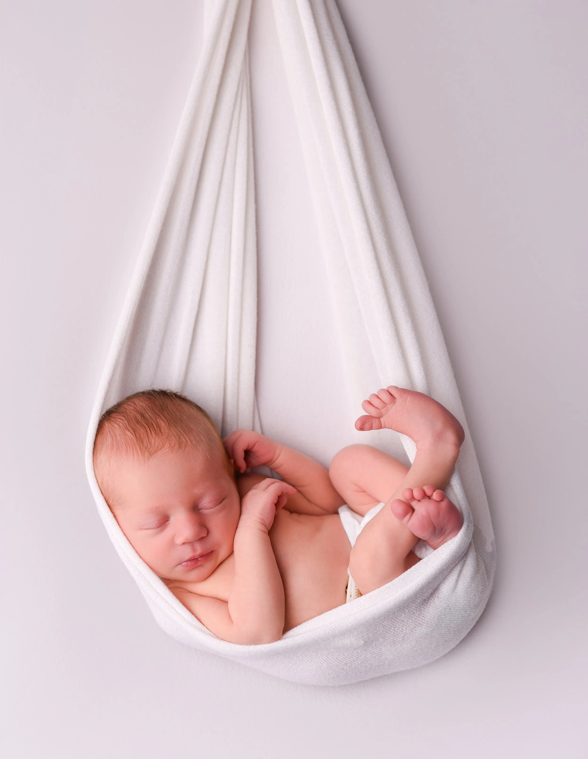 A newborn baby sleeping in a white hanging hammock against a plain white background.