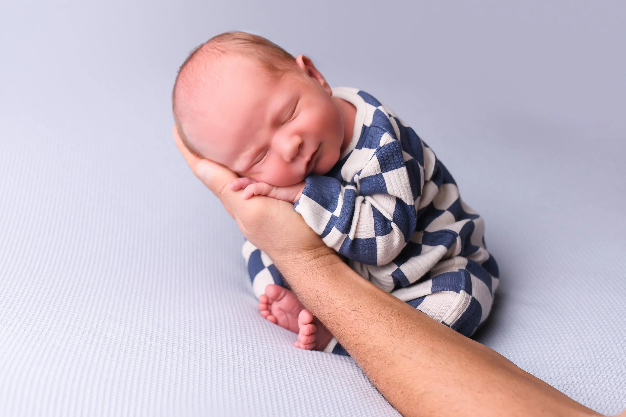 A newborn baby is peacefully sleeping on a light blue surface, cradled in an adult's hand, wearing a navy and white striped onesie.