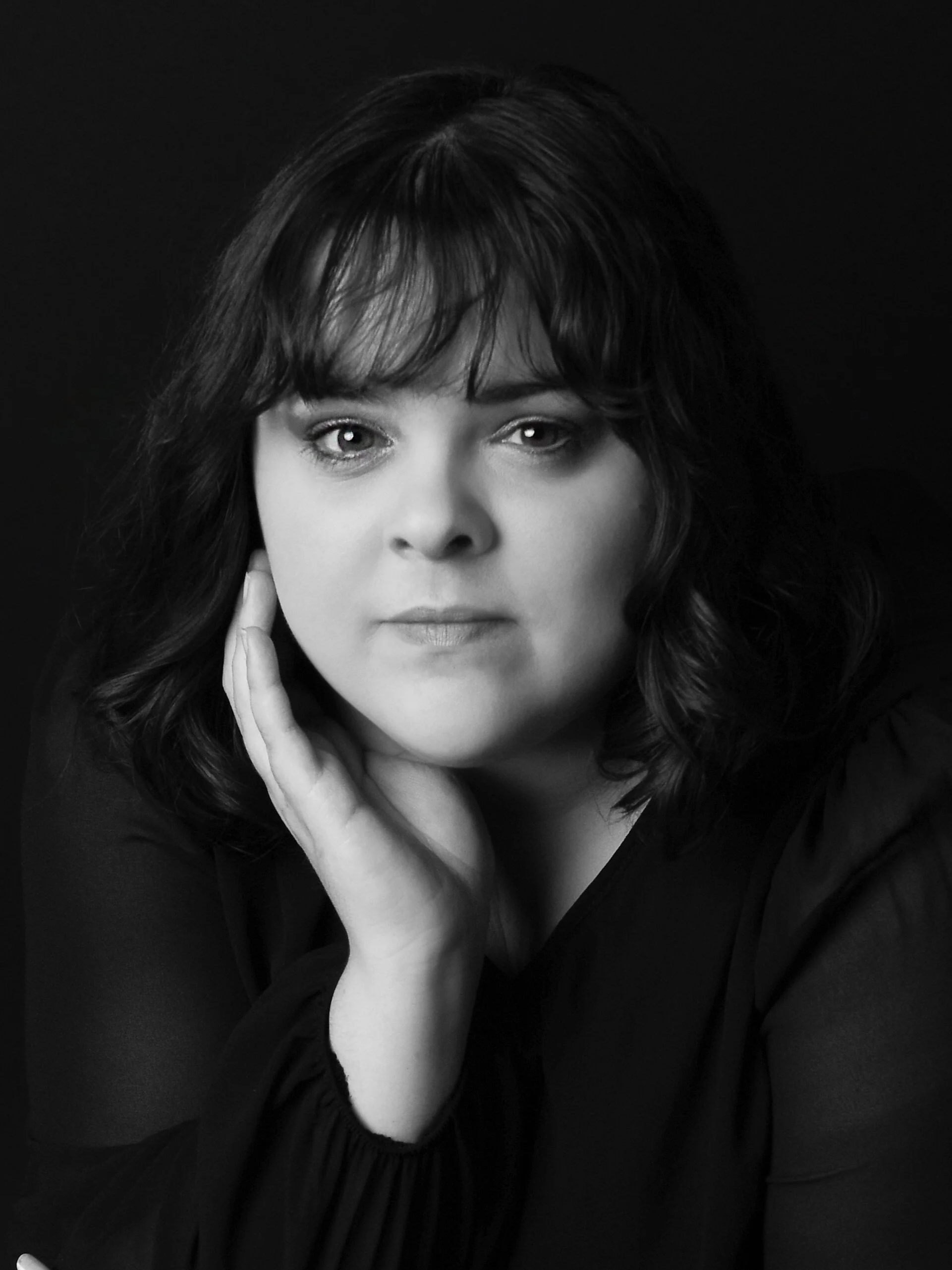 Black and white portrait of a woman with dark wavy hair, resting her chin on her hand, looking directly at the camera.