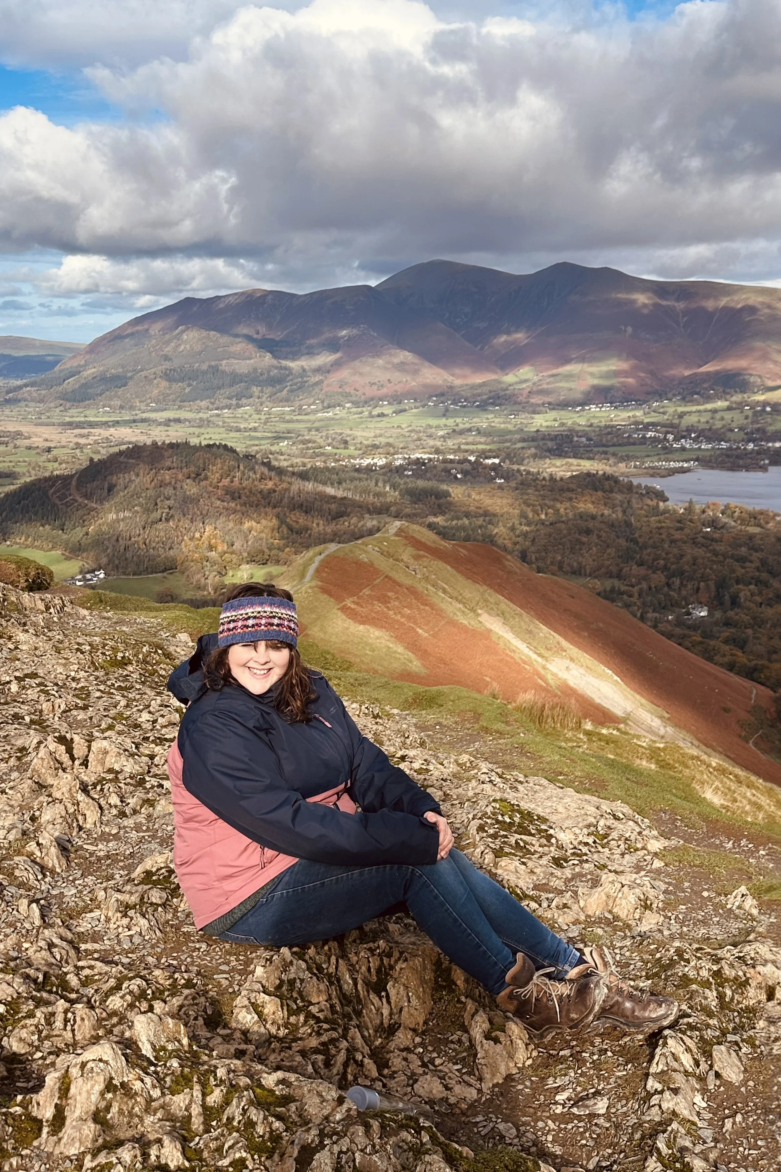 A woman sitting on rocky terrain on a mountain trail with a scenic view of rolling hills, a lake, and mountains under a partly cloudy sky.
