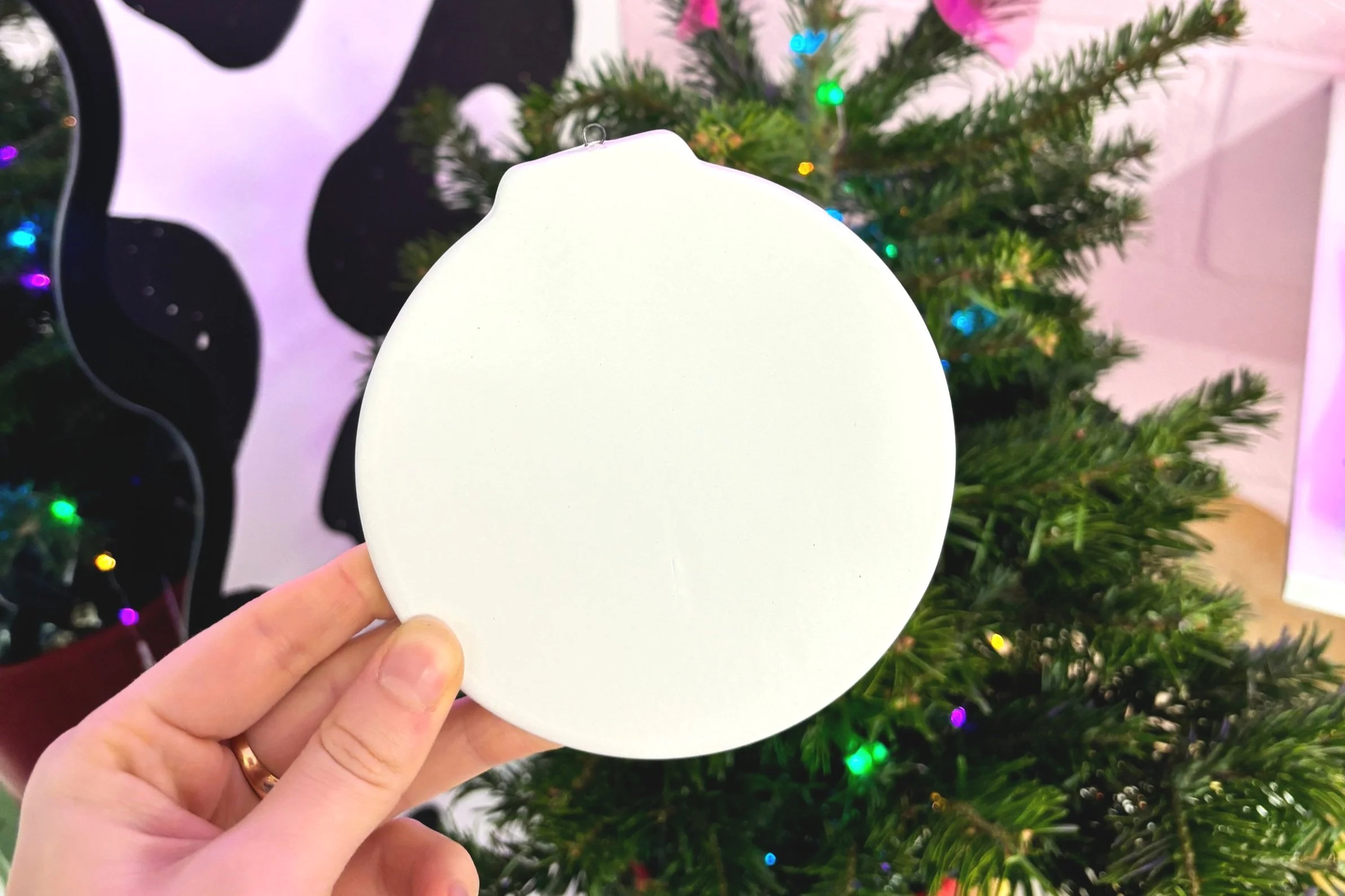 A hand holding a blank white ornament in front of a decorated Christmas tree with colorful lights.