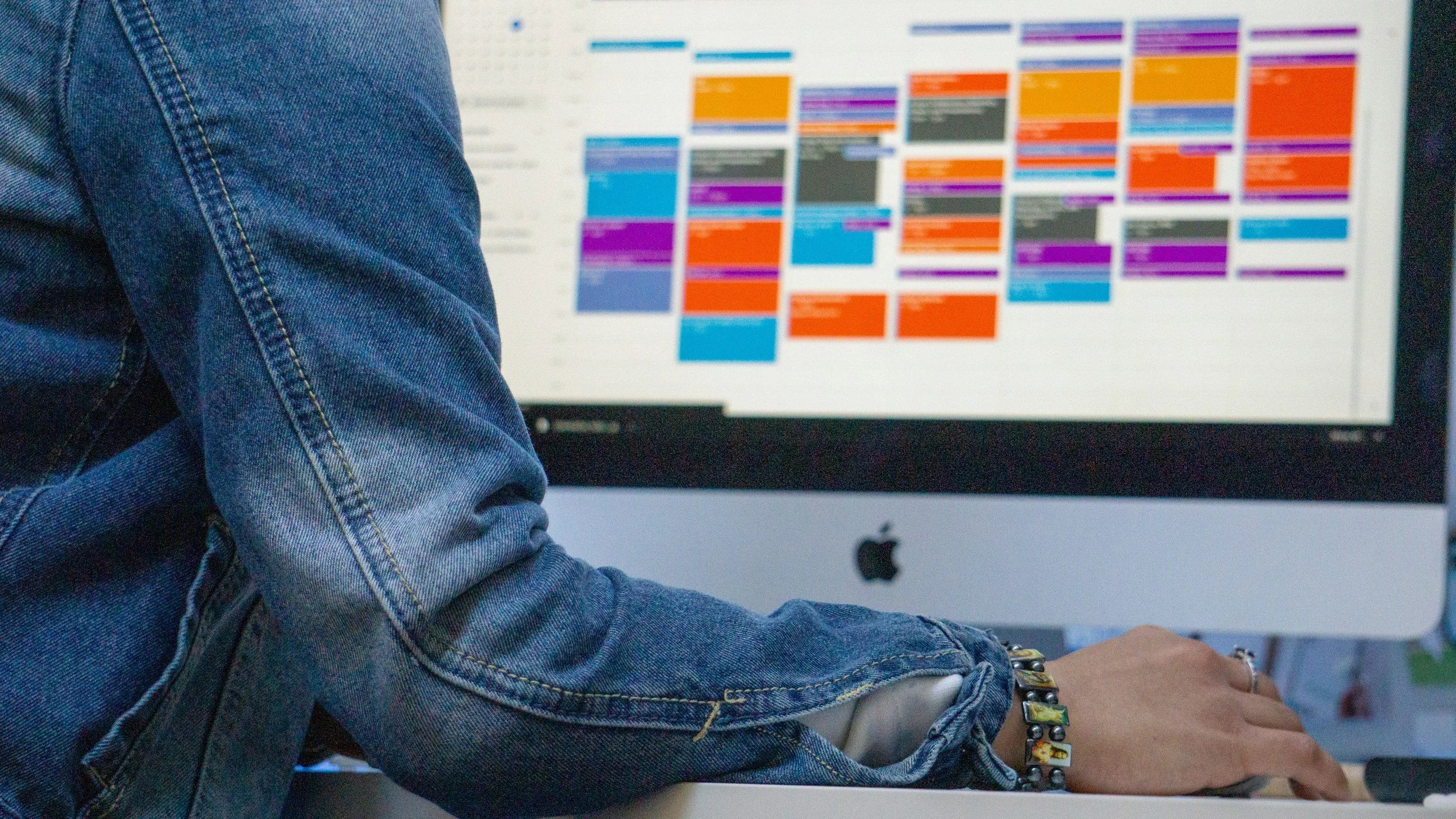 A person setting infront of an iMac with a full screen color-coded calendar.