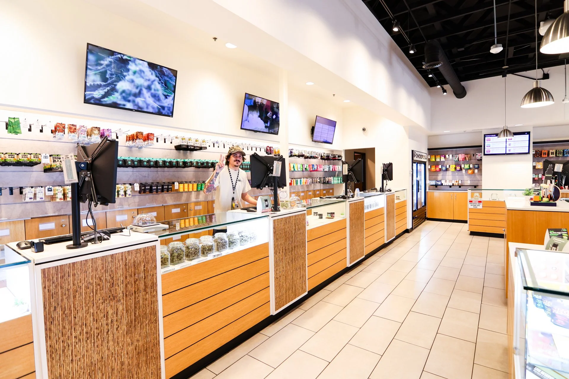 Inside a modern dispensary store with a friendly employee behind the counter giving a peace sign. Shelves with various products and multiple TV screens are visible, with a clean tiled floor and contemporary ceiling lights.