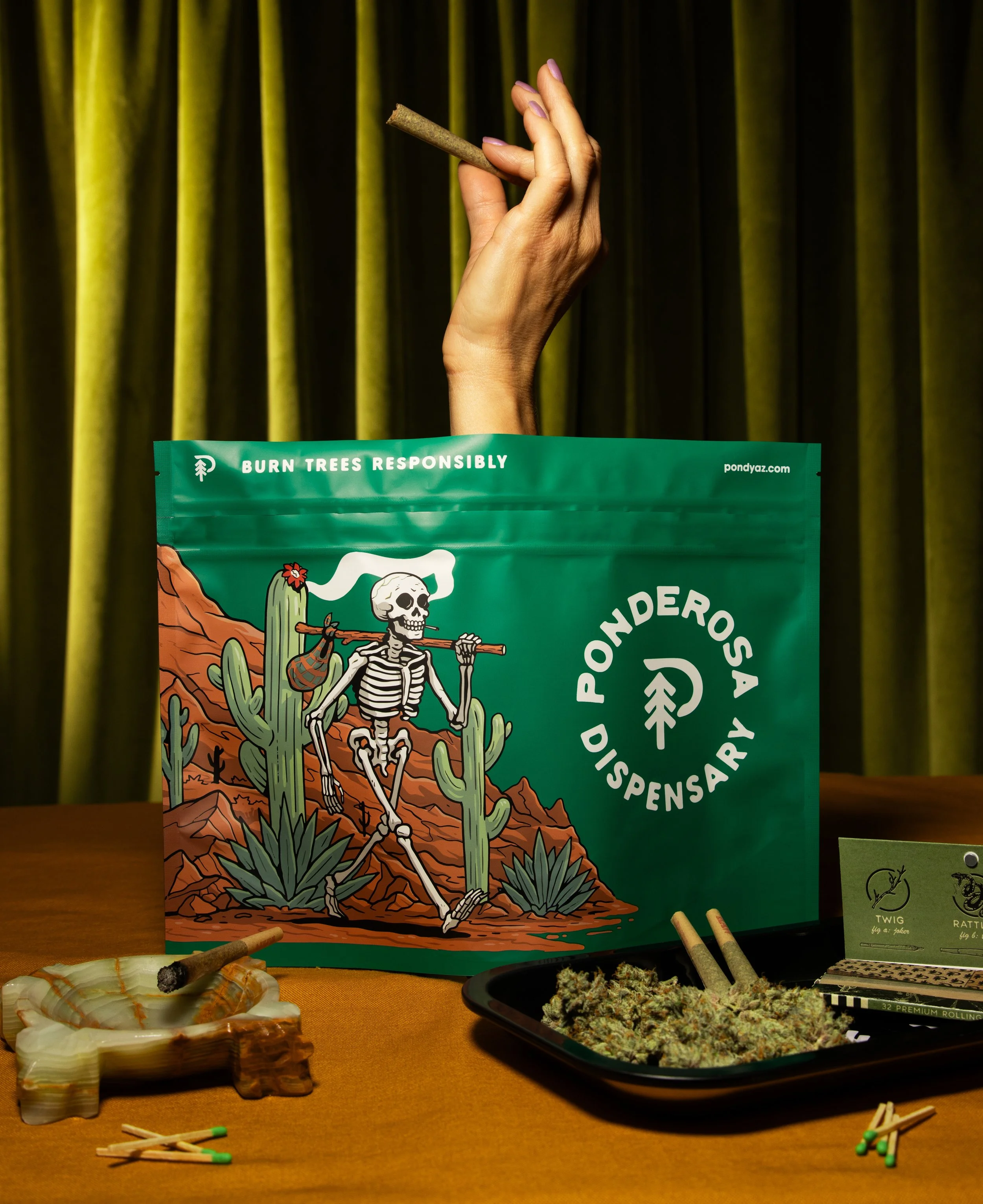 A poster featuring a skeleton in a desert scene with cactus, and text promoting responsible burning of trees. On the table, there are marijuana buds, matches, and rolling papers, with a background of yellow curtains.