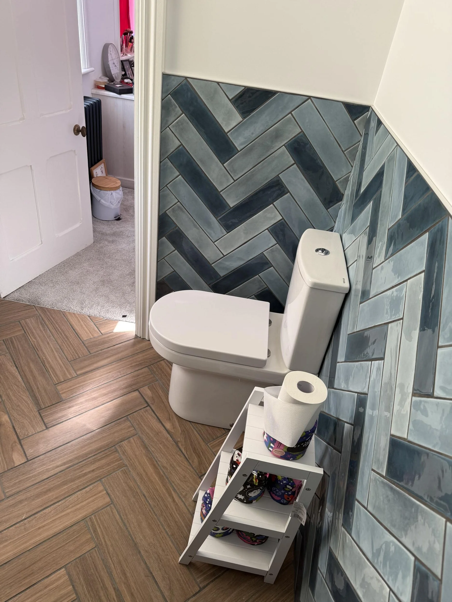 A bathroom with a toilet, a small white shelf holding a pack of toilet paper, a colorful cup, and some items that are not clearly visible. The wall behind the toilet is tiled with blue and gray tiles in a chevron pattern. Part of a multicolored room 