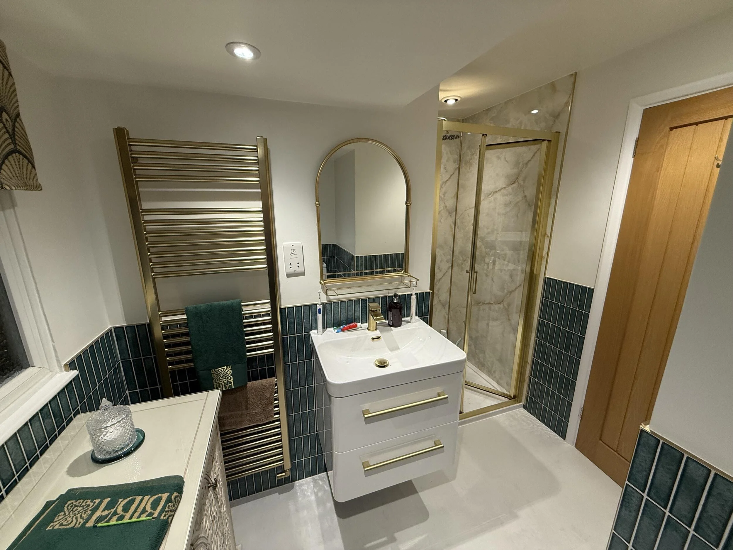 A bathroom with a gold towel warmer, a mirror, a white sink, and a walk-in shower with a glass door and marble walls.