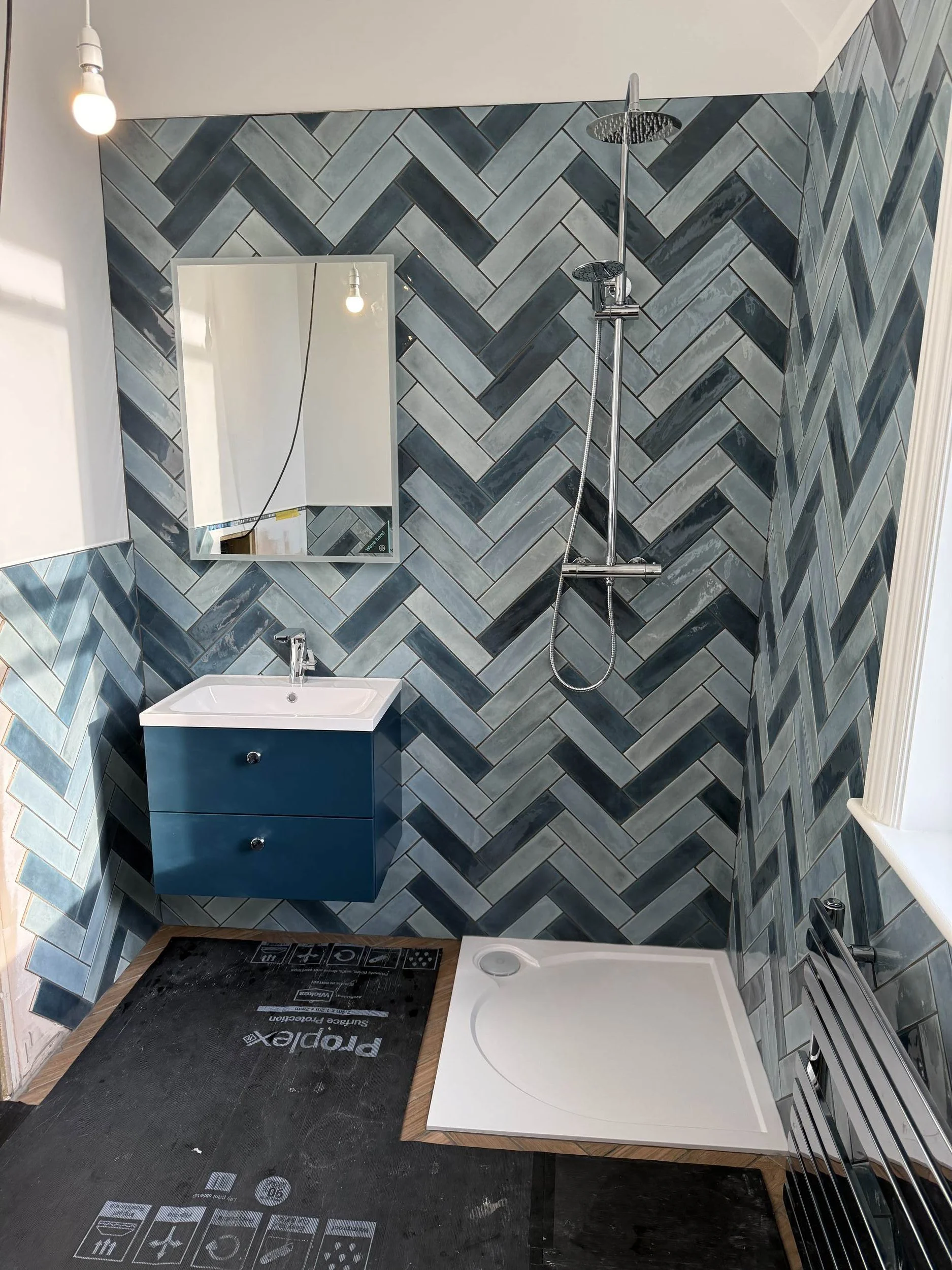A bathroom with dark herringbone tile walls, a mirror, a toilet, a sink with a blue cabinet, and a shower area with a rainfall showerhead.