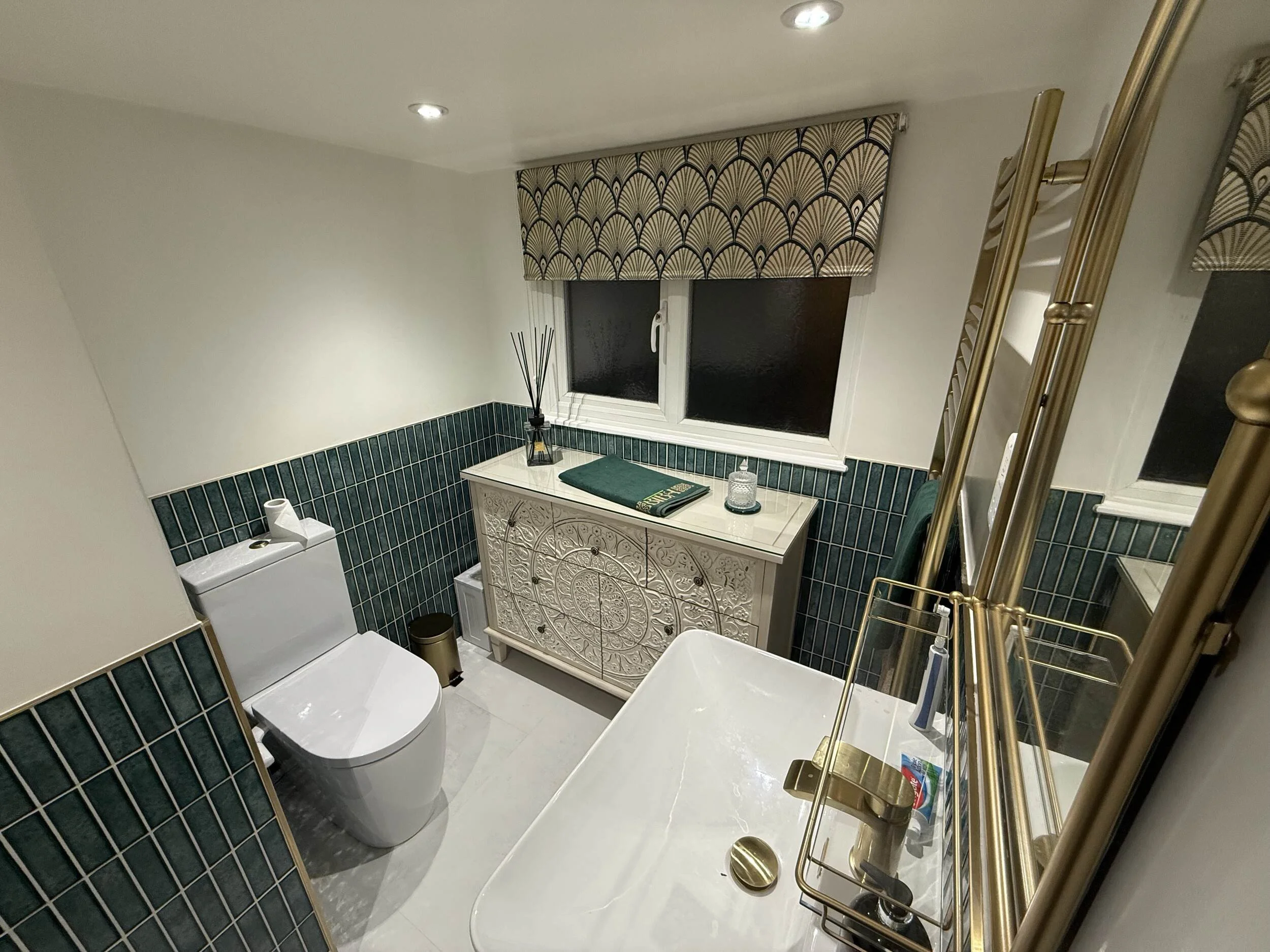 A small bathroom with a toilet, a decorative white cabinet, a mirror, a window with a patterned beige and black shade, green tile wainscoting, and gold fixtures including a towel rack and a ladder.