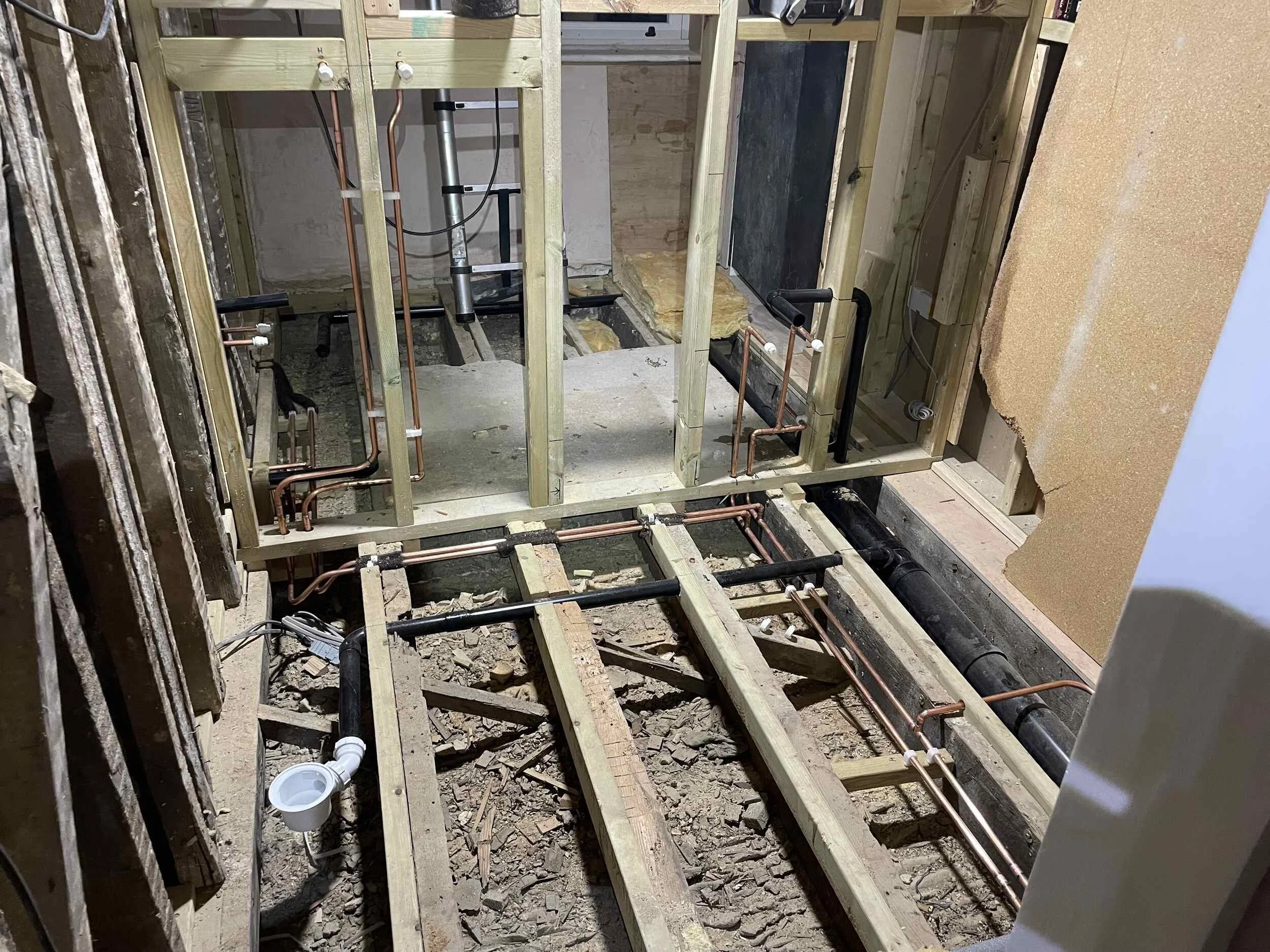 Interior view of a building under construction, showing exposed wooden framing, plumbing pipes, and insulation panels.