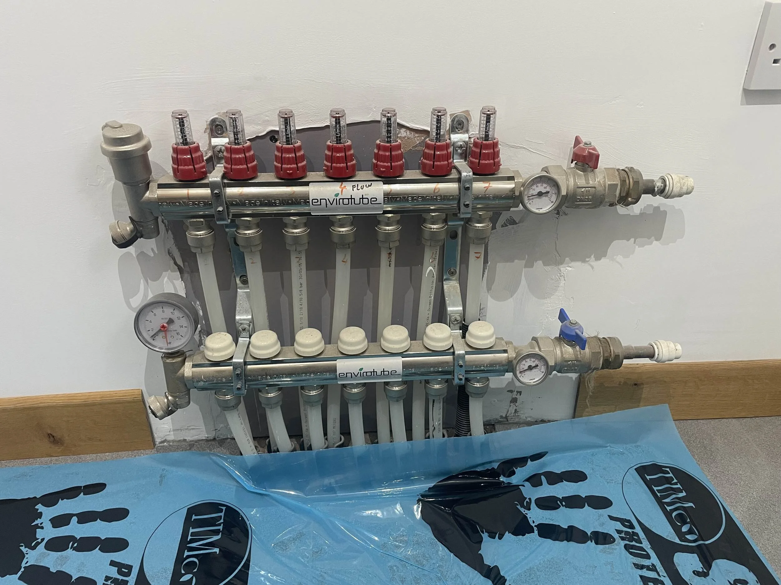 A plumbing manifold with multiple labeled valves and pressure gauges mounted on a wall, with a blue plastic sheet underneath.