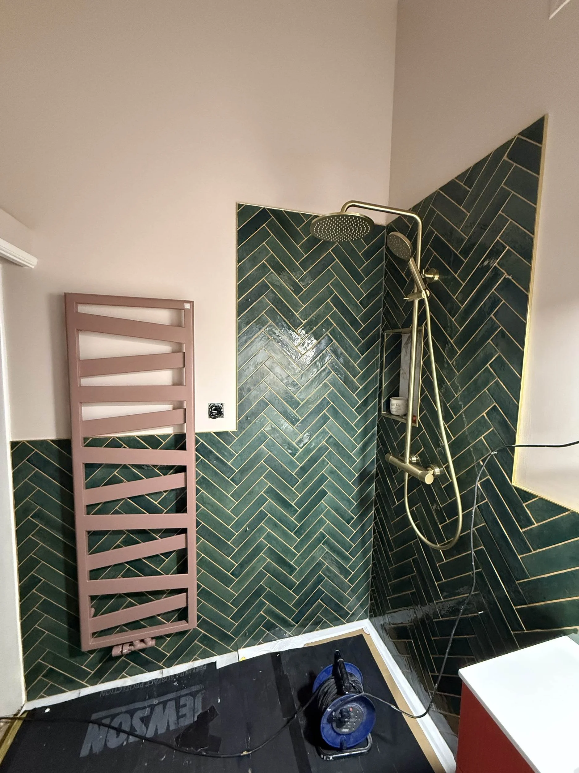 A bathroom shower area with dark green herringbone tile on the walls, a pink towel rack, and a black floor with a blue extension cord and a power drill on it. Part of a white wall and a red cabinet are also visible.