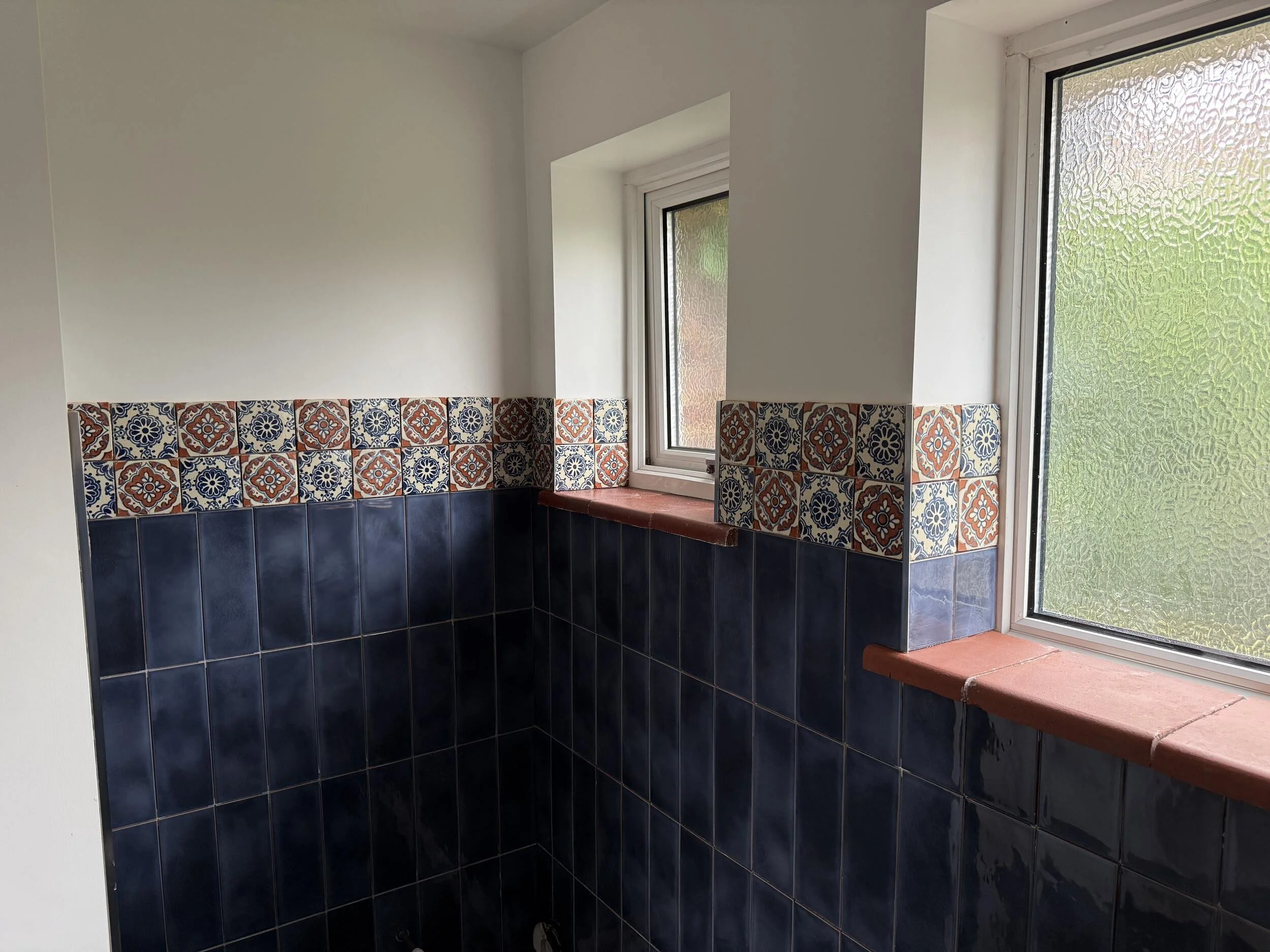 Interior of a room with white walls, decorative patterned tiles in red, blue, and white, a window and glass door with textured glass, and a red brick ledge.
