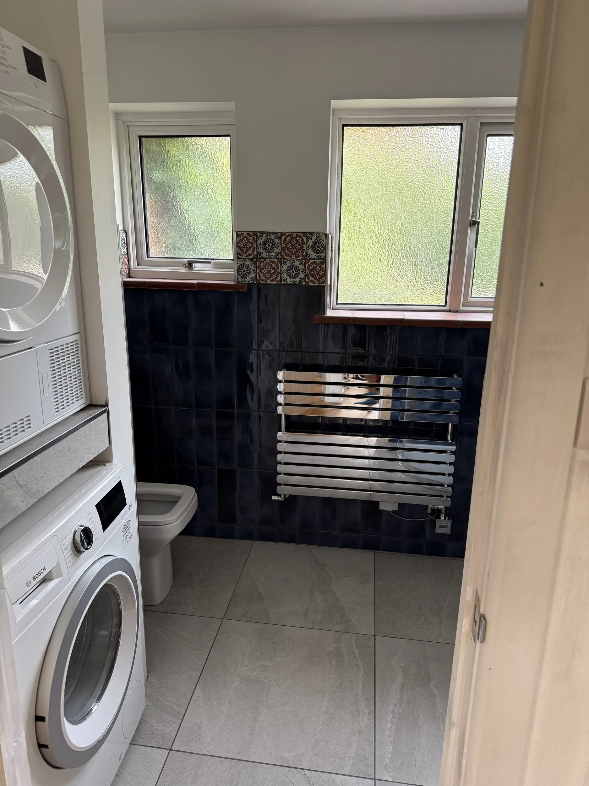 A laundry room with front-loading washing machine and tumble dryer on the left, a small toilet in the corner, two windows with frosted glass, dark blue tiles, a wooden shelf, and a wall-mounted heated towel rail.