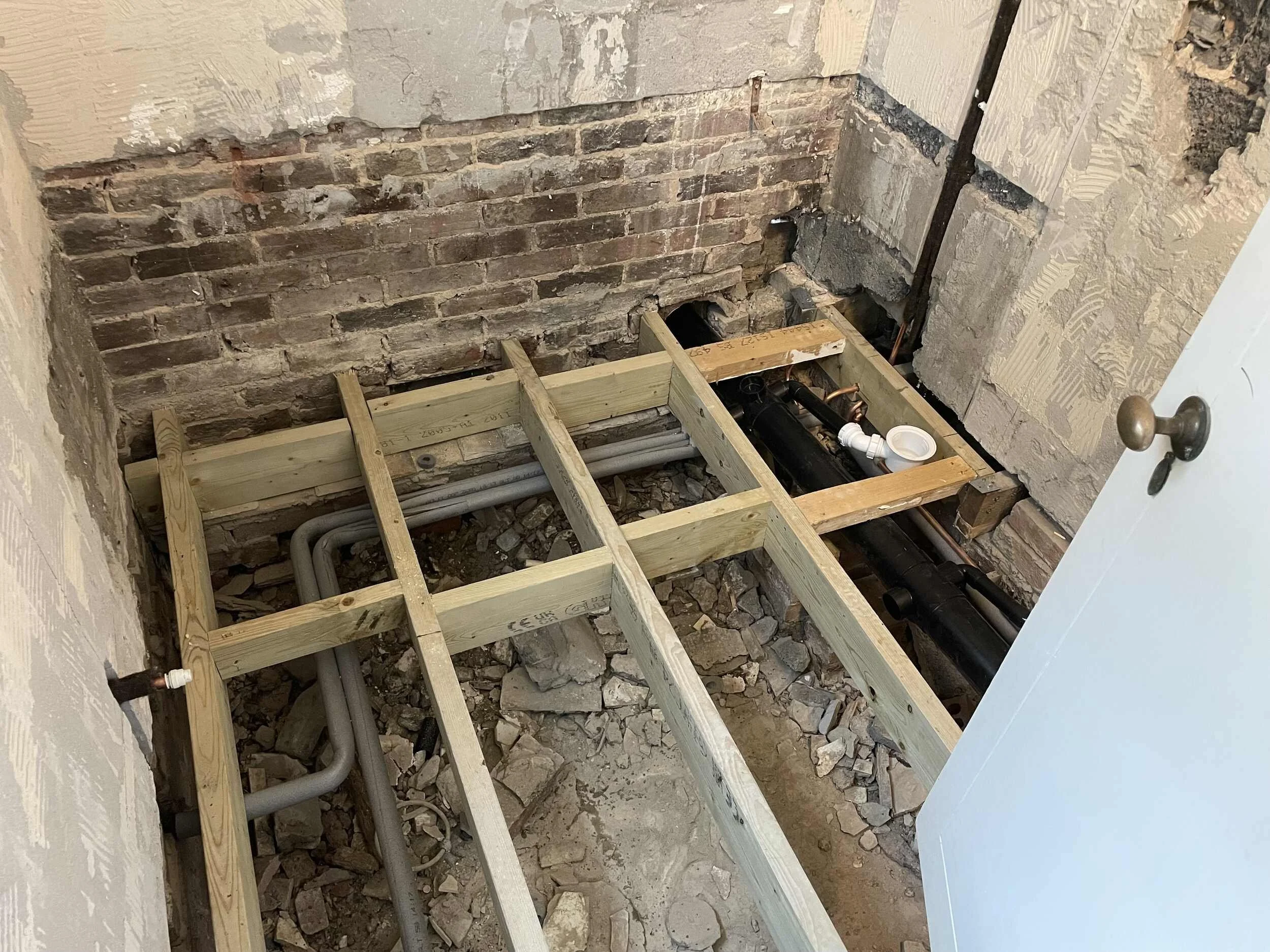 Construction site with exposed brick wall, wooden framing, pipes, and debris on the floor, indicating ongoing renovation or plumbing work.