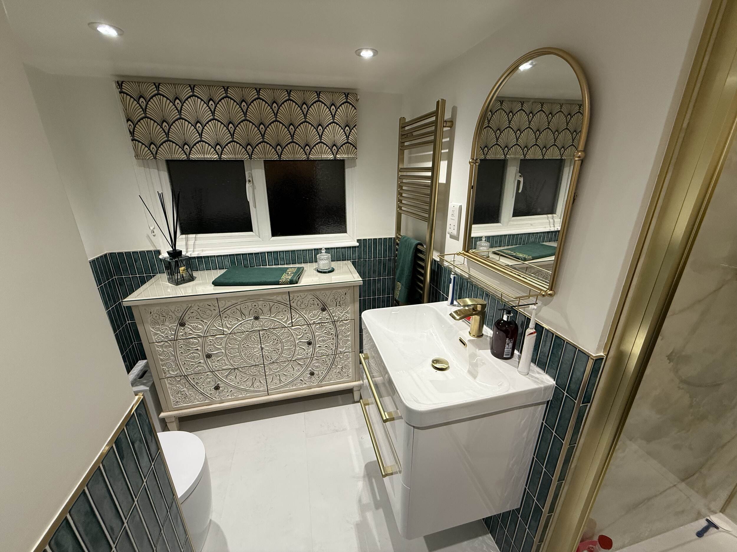A bathroom with a white ornate vanity, a small sink, a gold-framed mirror, blue tiles, and a green towel. There is a window with a patterned curtain and a basket with glass bottles on the vanity.