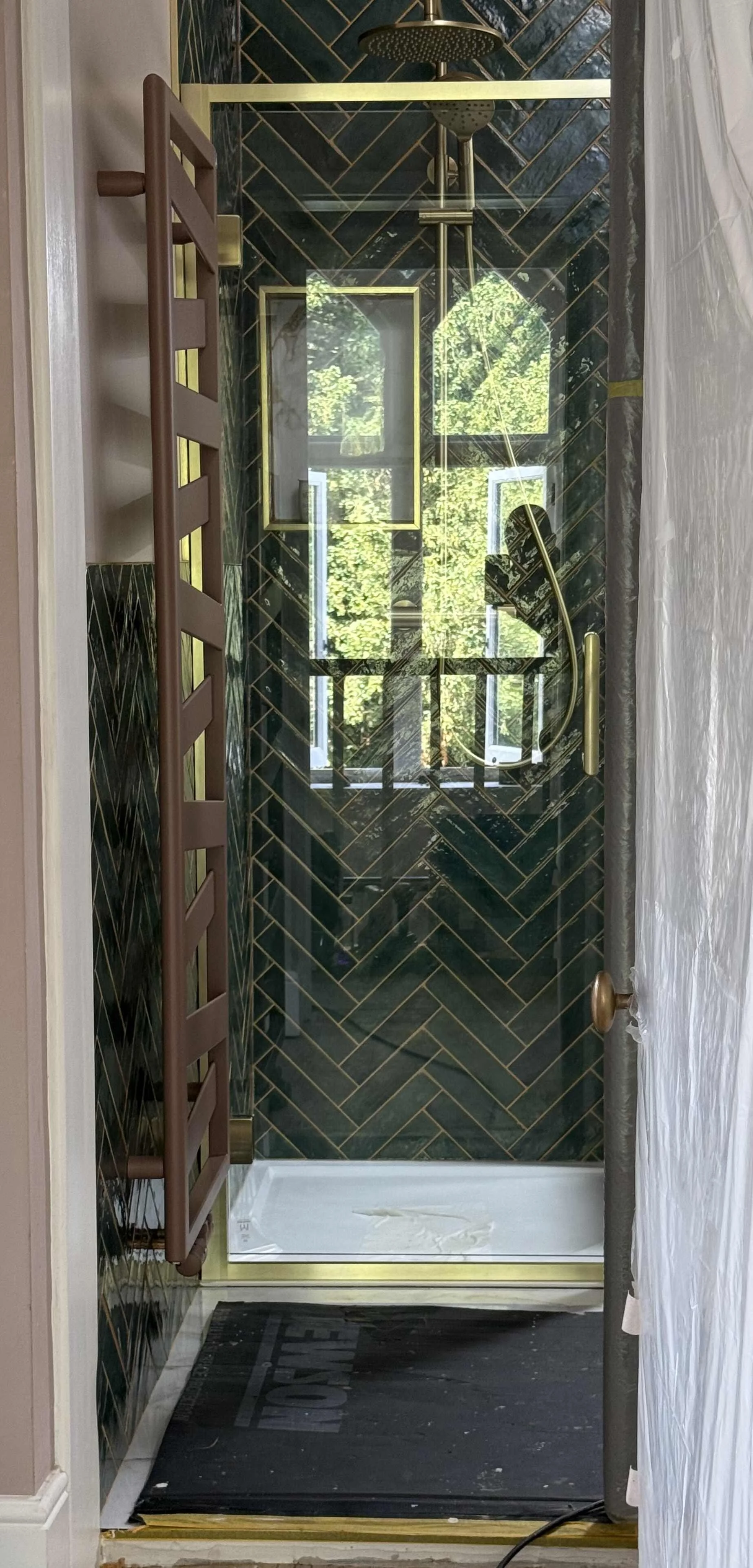 A modern bathroom shower with black tile arranged in a herringbone pattern, gold accents, and a metal rainfall showerhead. There is a window reflected in the glass with trees outside, and a brown towel rack on the left. The shower floor is white, and