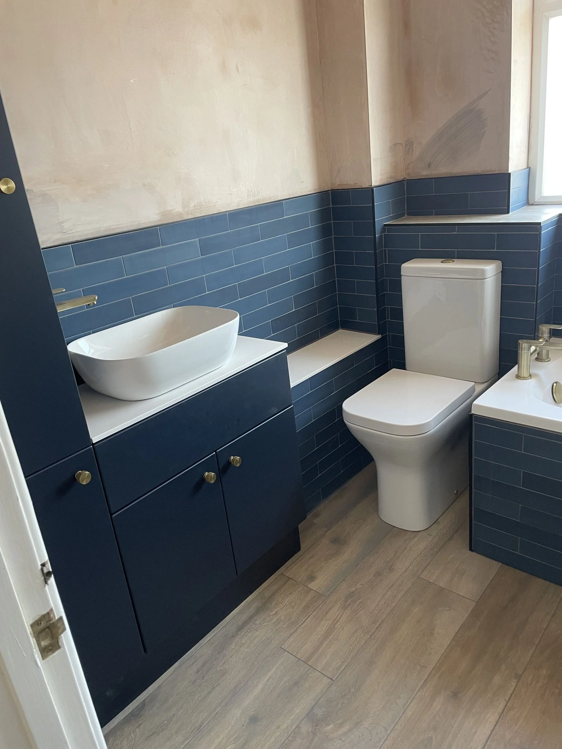 Modern bathroom with navy blue cabinets, a white vessel sink, a toilet, and natural light coming through a window, with wood-look flooring.