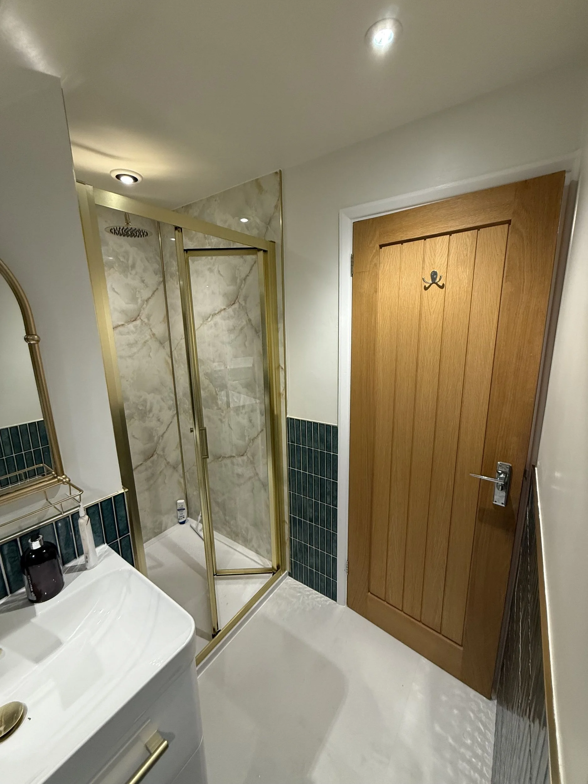 A small bathroom with a wooden door and a shower with glass sliding doors, beige marble walls, a white sink, and a mirror.