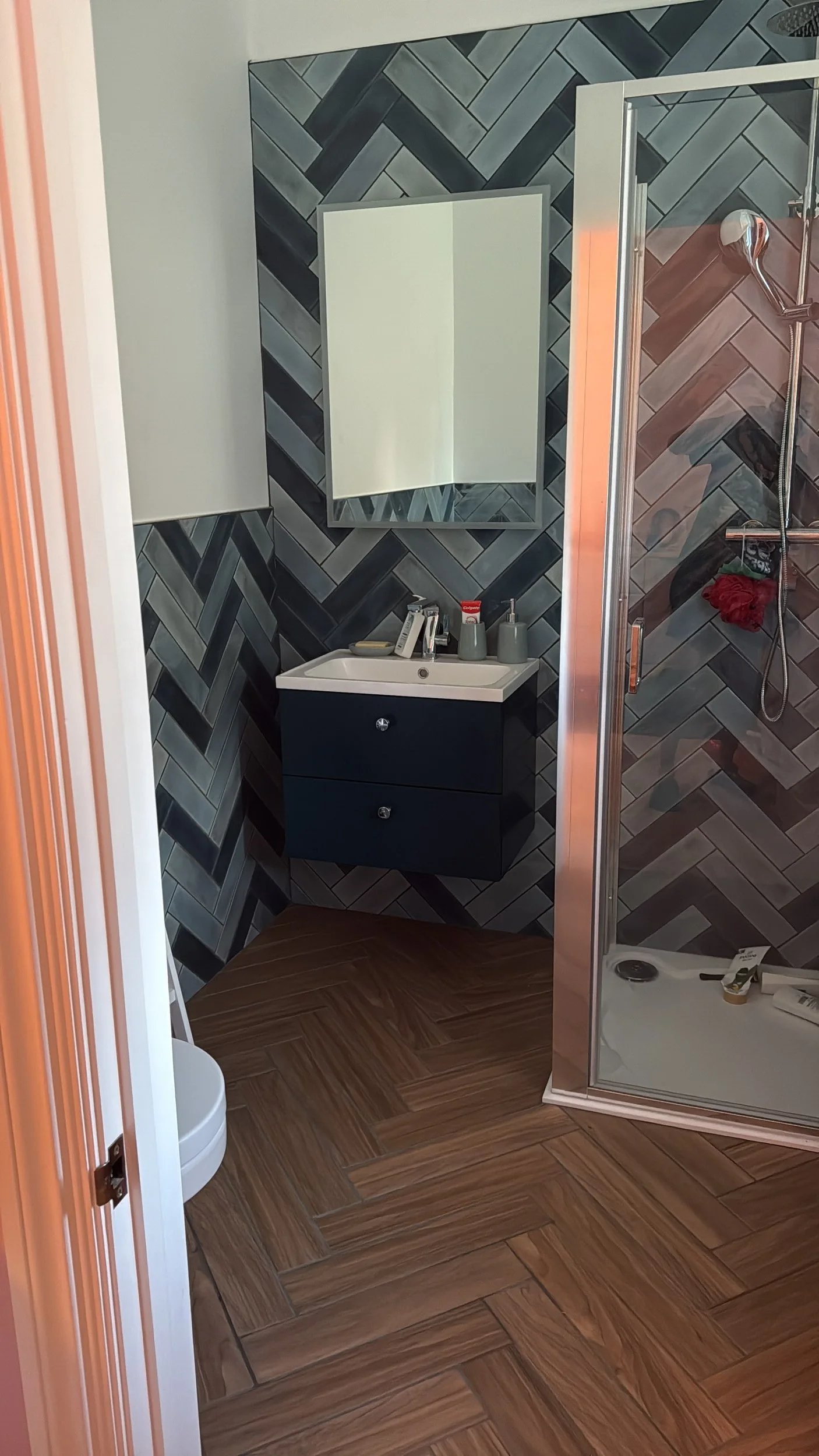 A small bathroom with a black vanity, a mirror, and a glass shower stall. The walls are decorated with gray and black herringbone tiles, and the flooring is wood-patterned tile.