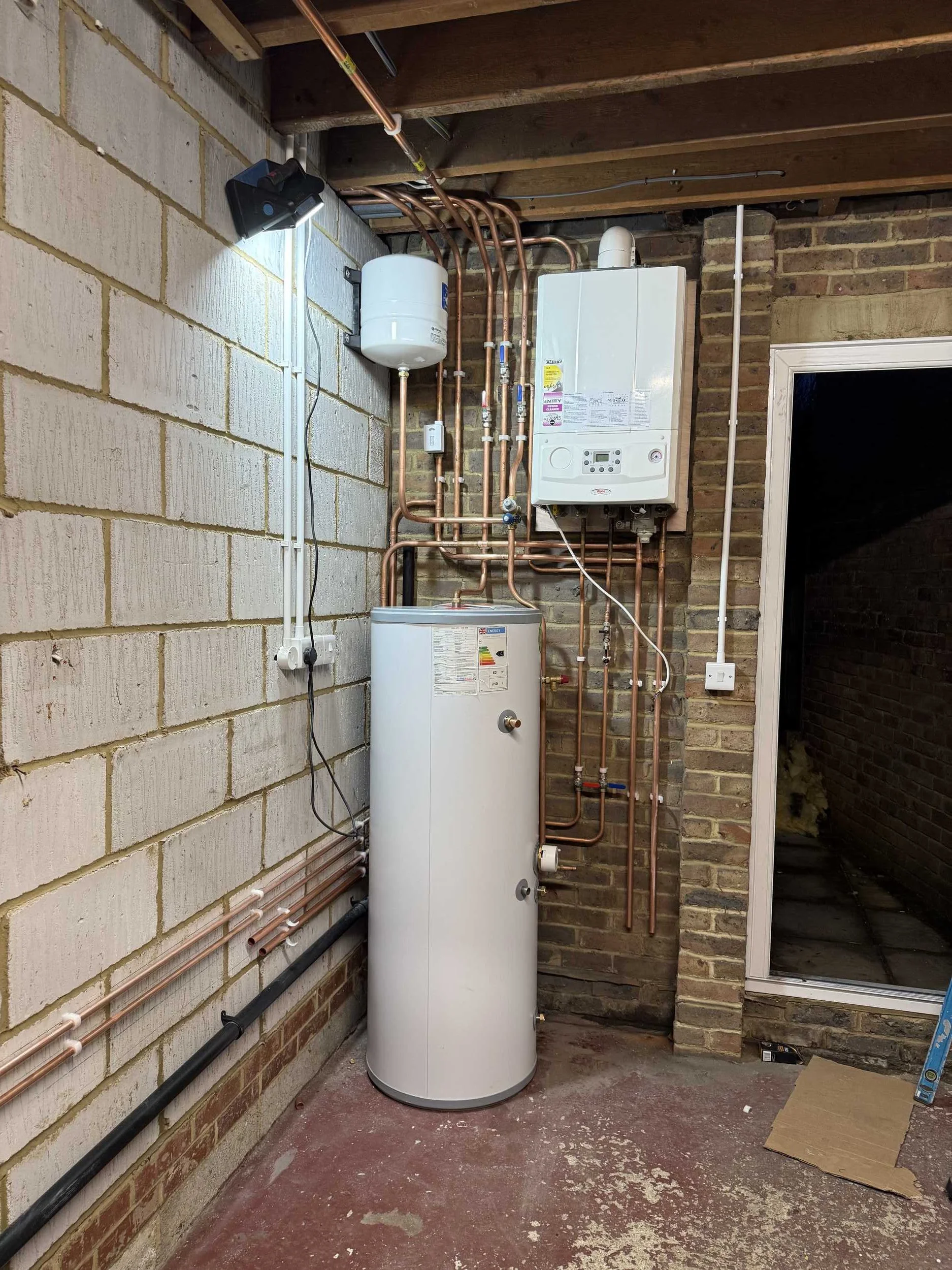 A utility room with exposed brick and concrete walls. Contains a white water heater, a white boiler, copper pipes, and electrical wiring. There is a window on the right and some tools on the floor.