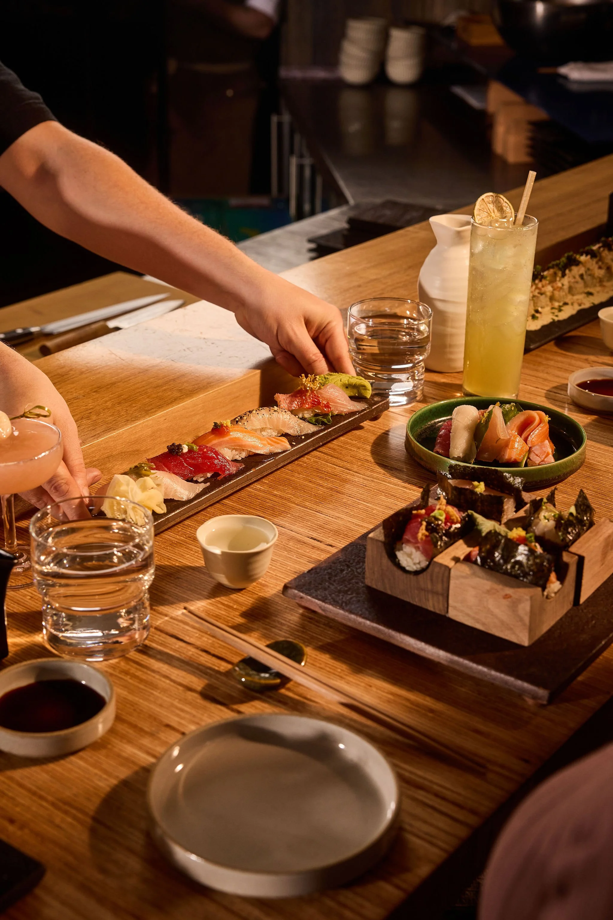 Sushi plates and drinks on a wooden table in a dimly lit restaurant.