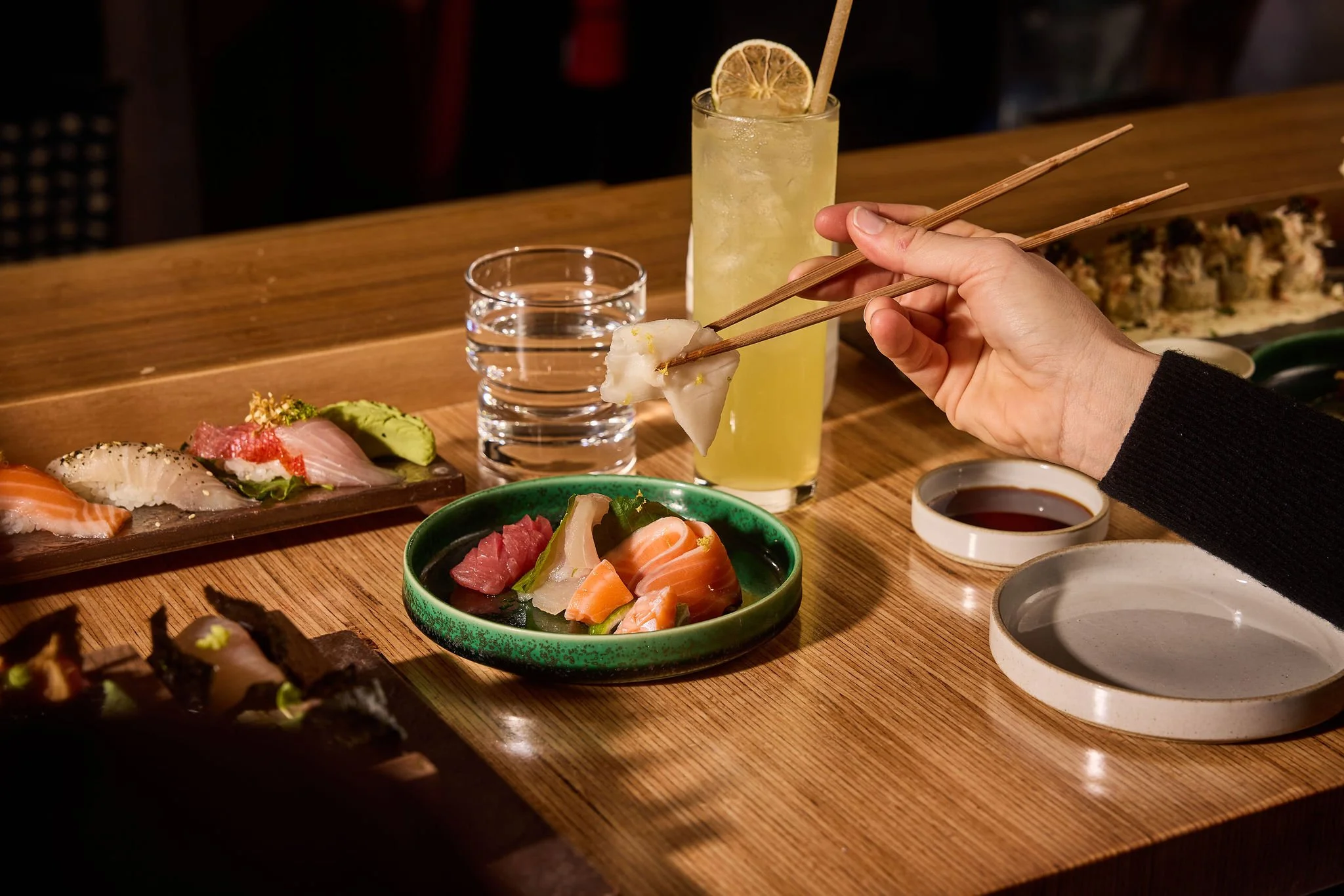 A person uses chopsticks to pick up a piece of sushi at a wooden table with various sushi, sashimi, a green ceramic bowl of sashimi, a tall glass of lemonade with a lemon slice, two glasses of water, soy sauce, and a small dish of dipping sauce.